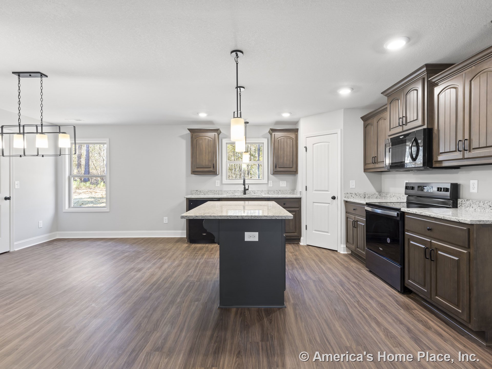 Open kitchen with wood cabinetry, granite countertops, central island topped with pendant lights, recessed ceiling lighting, vinyl plank flooring, stainless steel microwave and