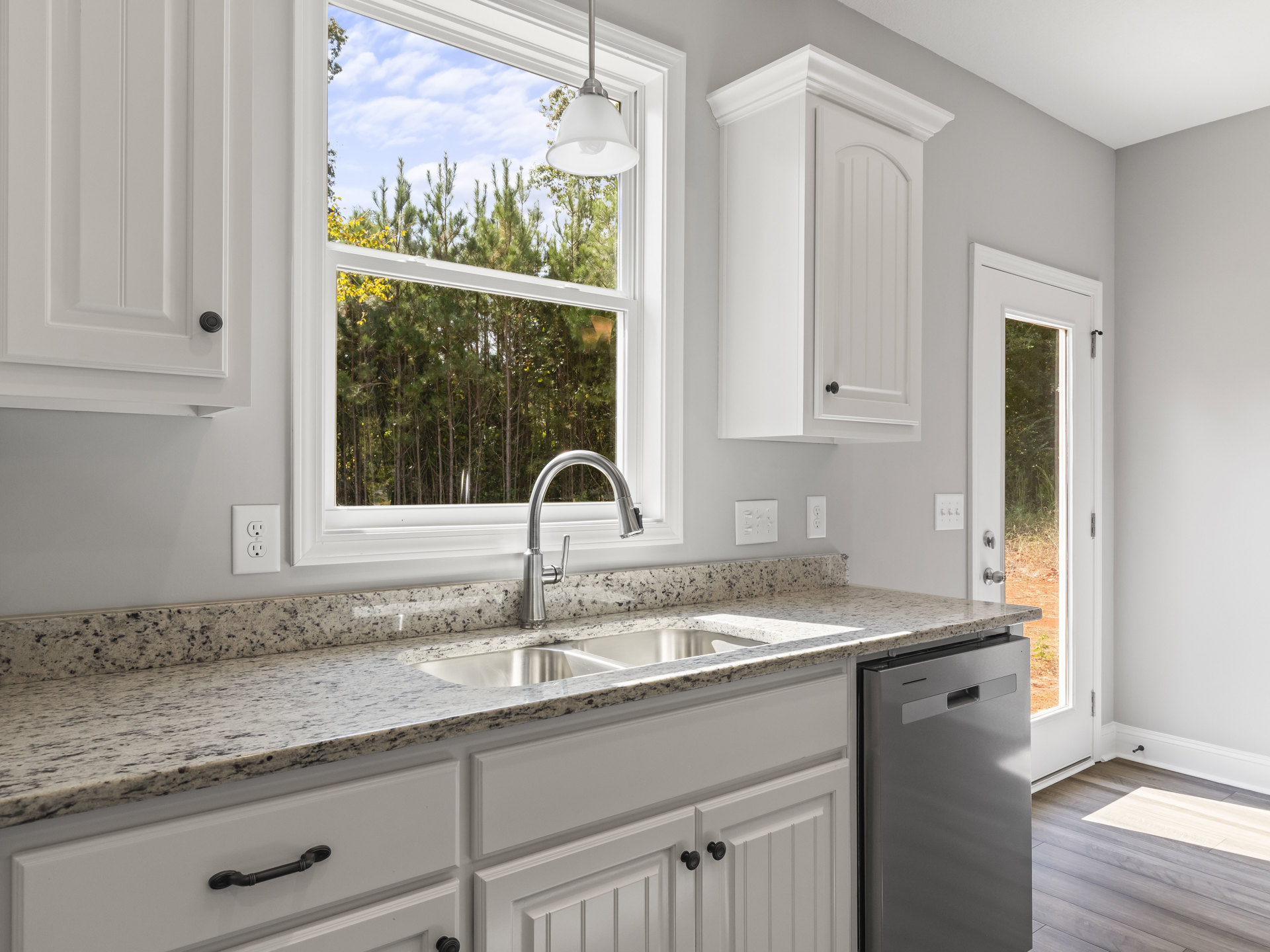 Modern kitchen featuring white tile backsplash, stainless steel sink and dishwasher, shaker-style cabinetry, stone countertop, chrome faucet, large window with natural light, and