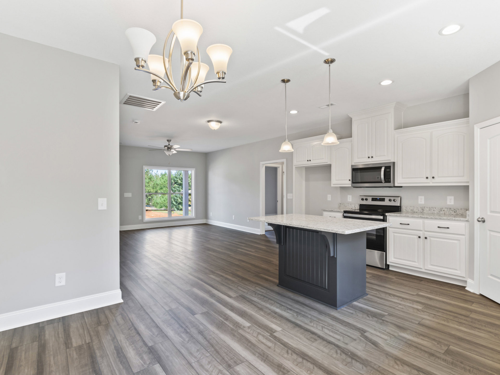 Kitchen and dining room with wood flooring, marble-topped island, open microwave, white-shaded light fixture, cabinetry, and window showing trees outside