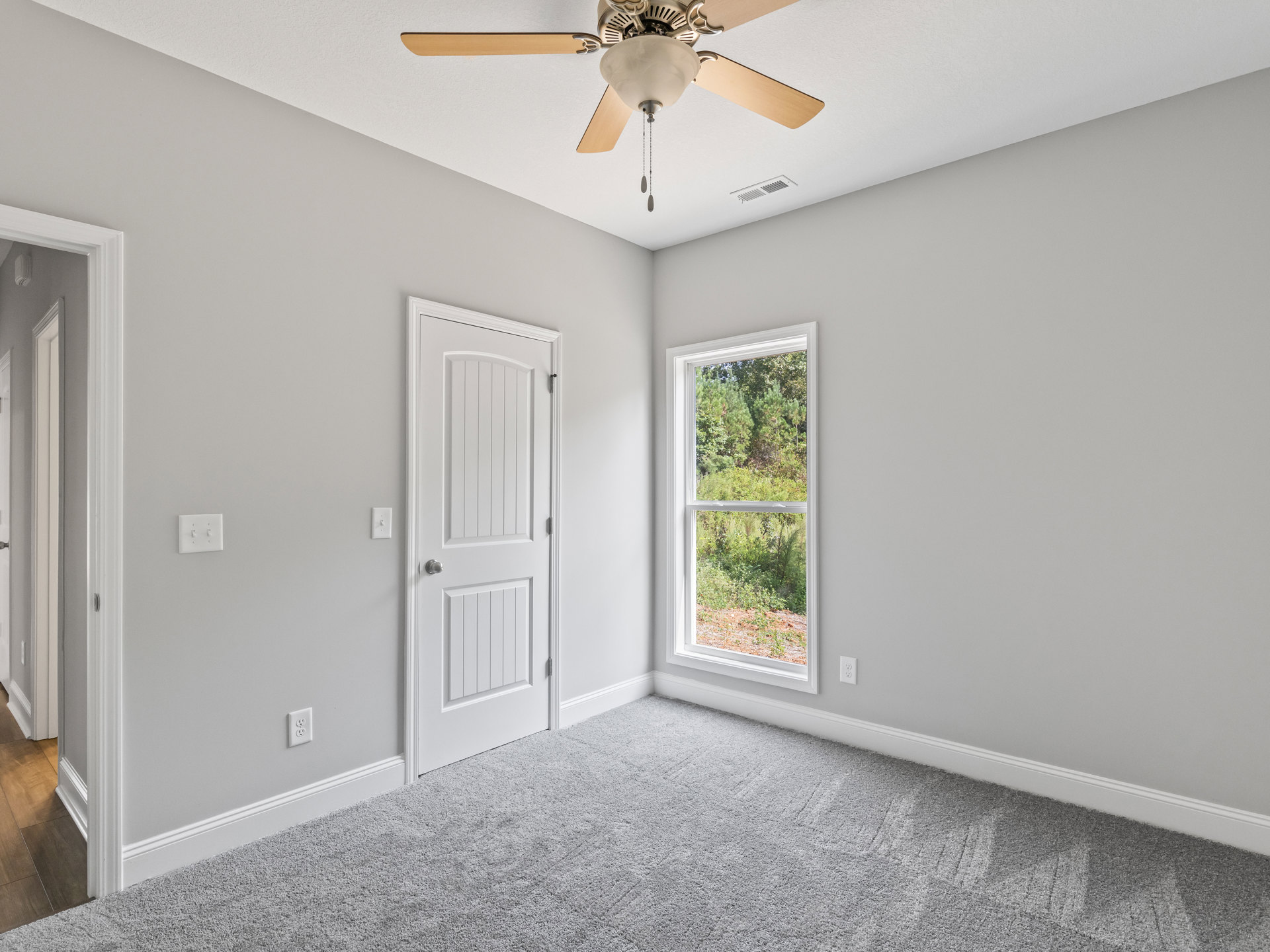 Carpeted room with white walls, ceiling fan with light fixture, white door with silver knob, window showing trees outside, light switch on wall, simple baseboard molding