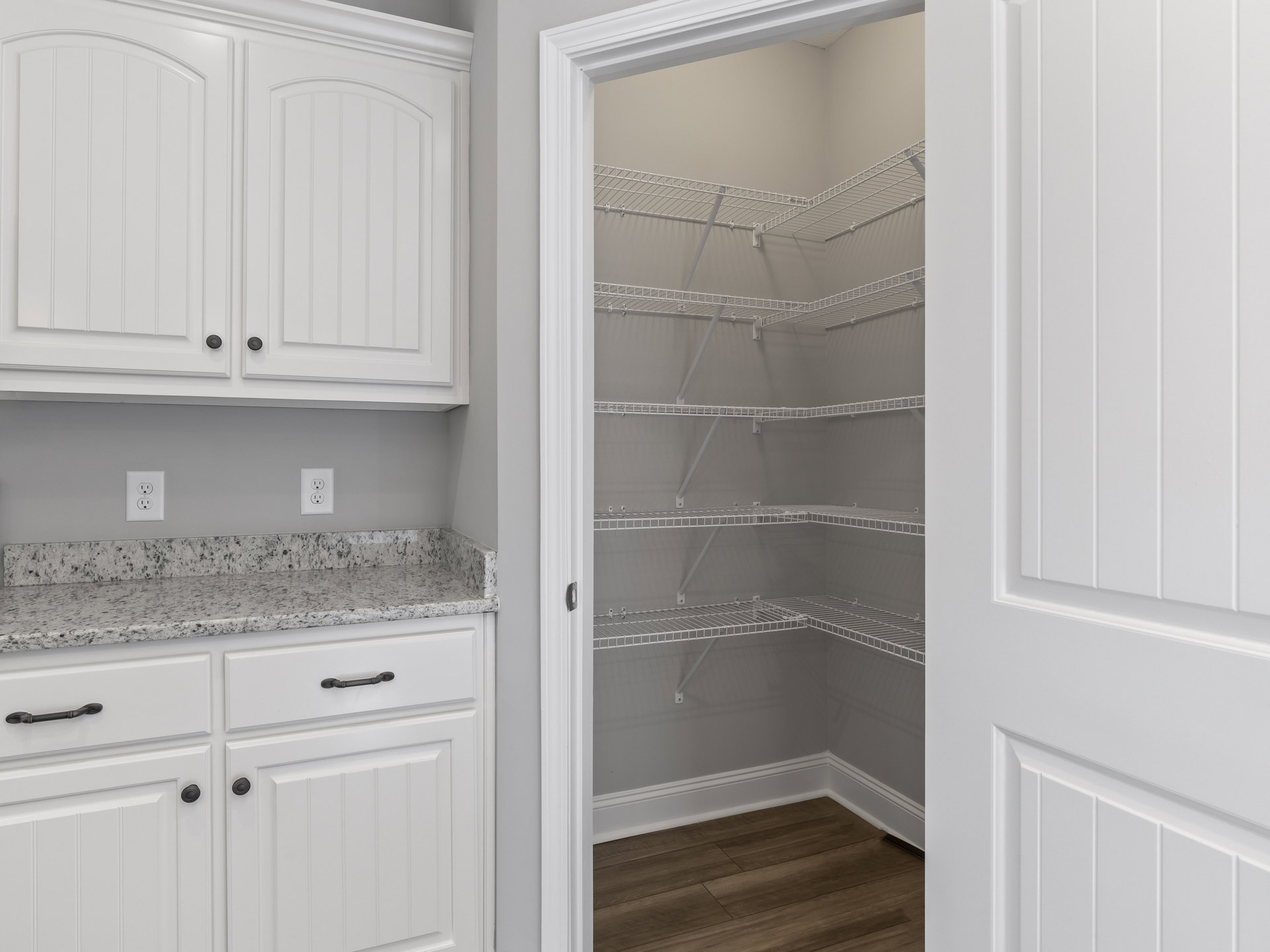 White closet with built-in shelves, wood floor and white baseboard, white cabinets with black knobs, curved-top white door, close-up of electrical outlet