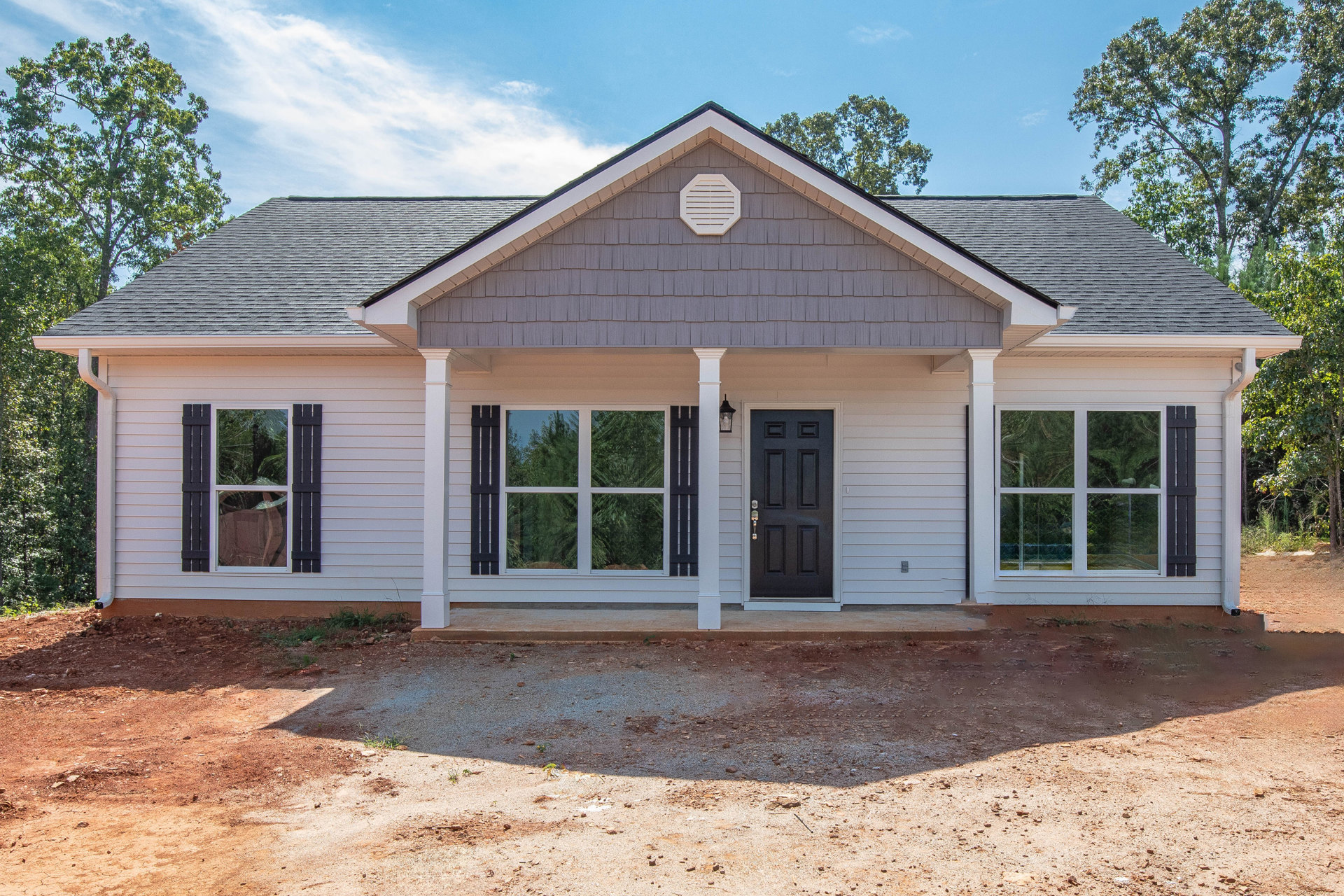Gray exterior home with black front door and silver handle, white vent on wall, driveway with dirt surface, large window reflecting trees and sky.