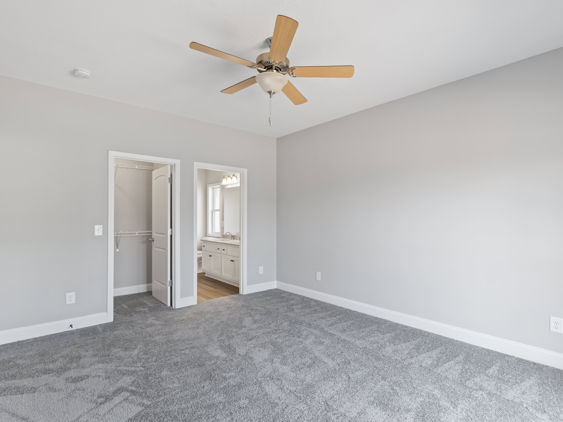 Bedroom with grey carpet, white closet door, ceiling fan with light fixture, and partial view of bathroom featuring sink and window