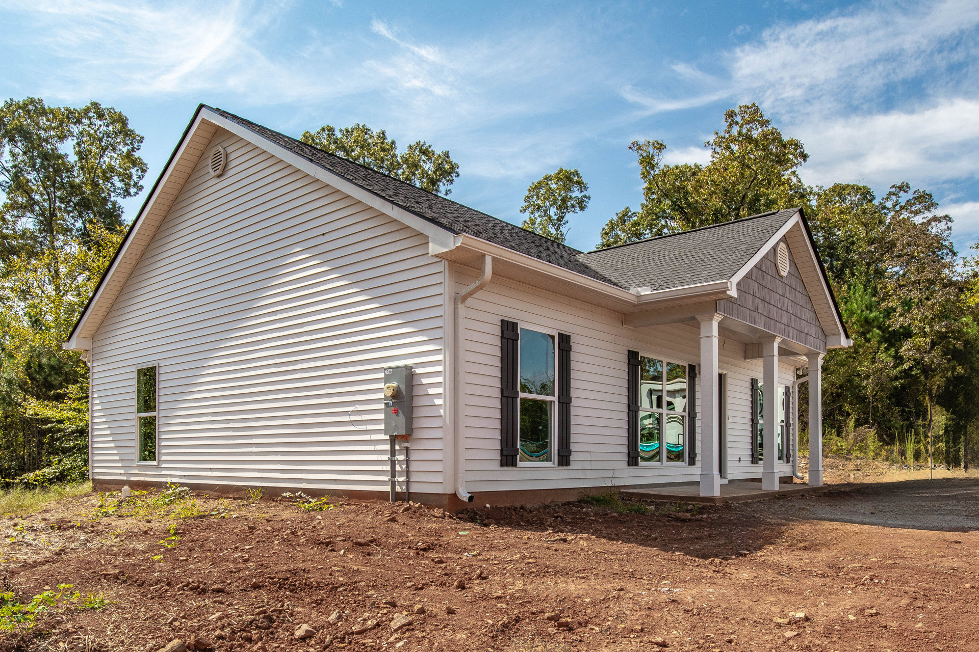 Modern cottage-style home surrounded by mature trees, unfinished dirt landscaping, large windows reflecting greenery and interior furnishings, visible utility meter on exterior