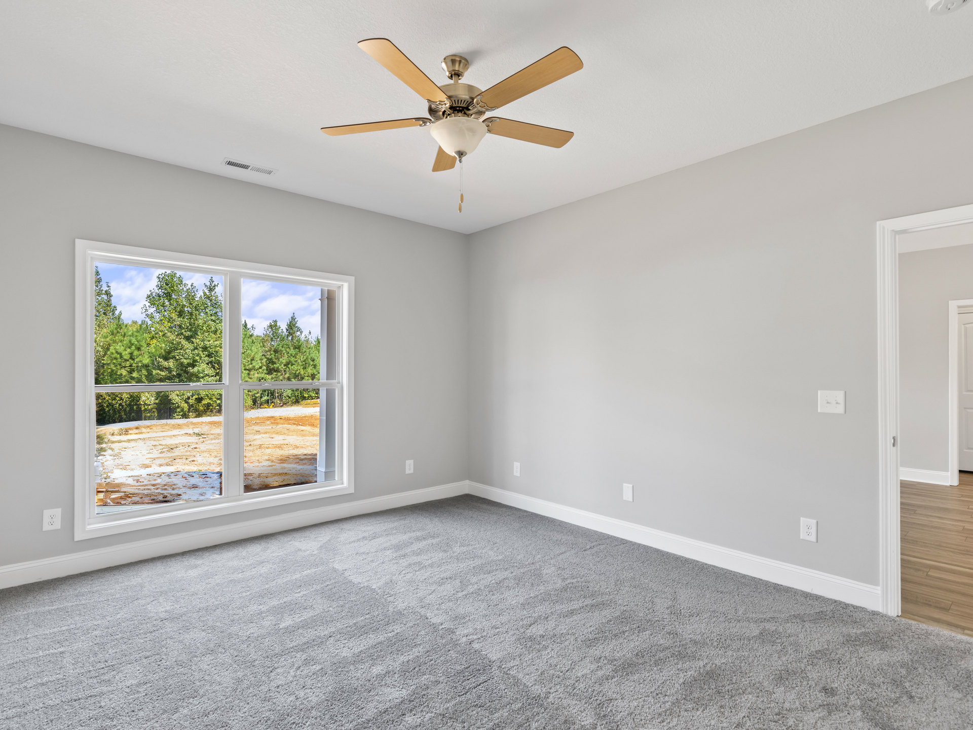 Ceiling fan with light fixture above grey carpeted floor, single window with view of trees and dirt field, white walls with crown molding