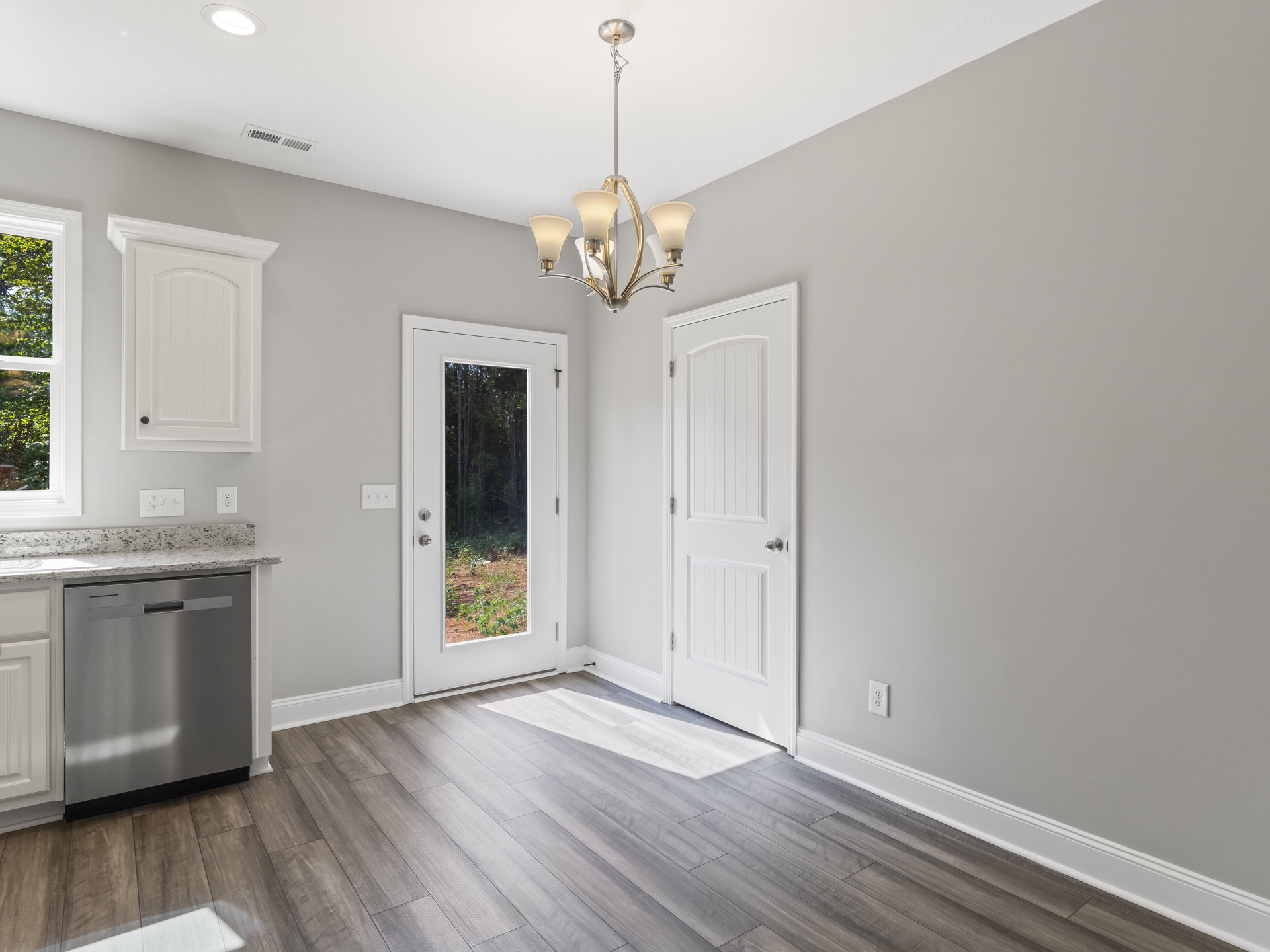 Open kitchen with white cabinetry, stainless steel dishwasher, laminate flooring, white door with silver handle, window overlooking forest, modern chandelier hanging from ceiling