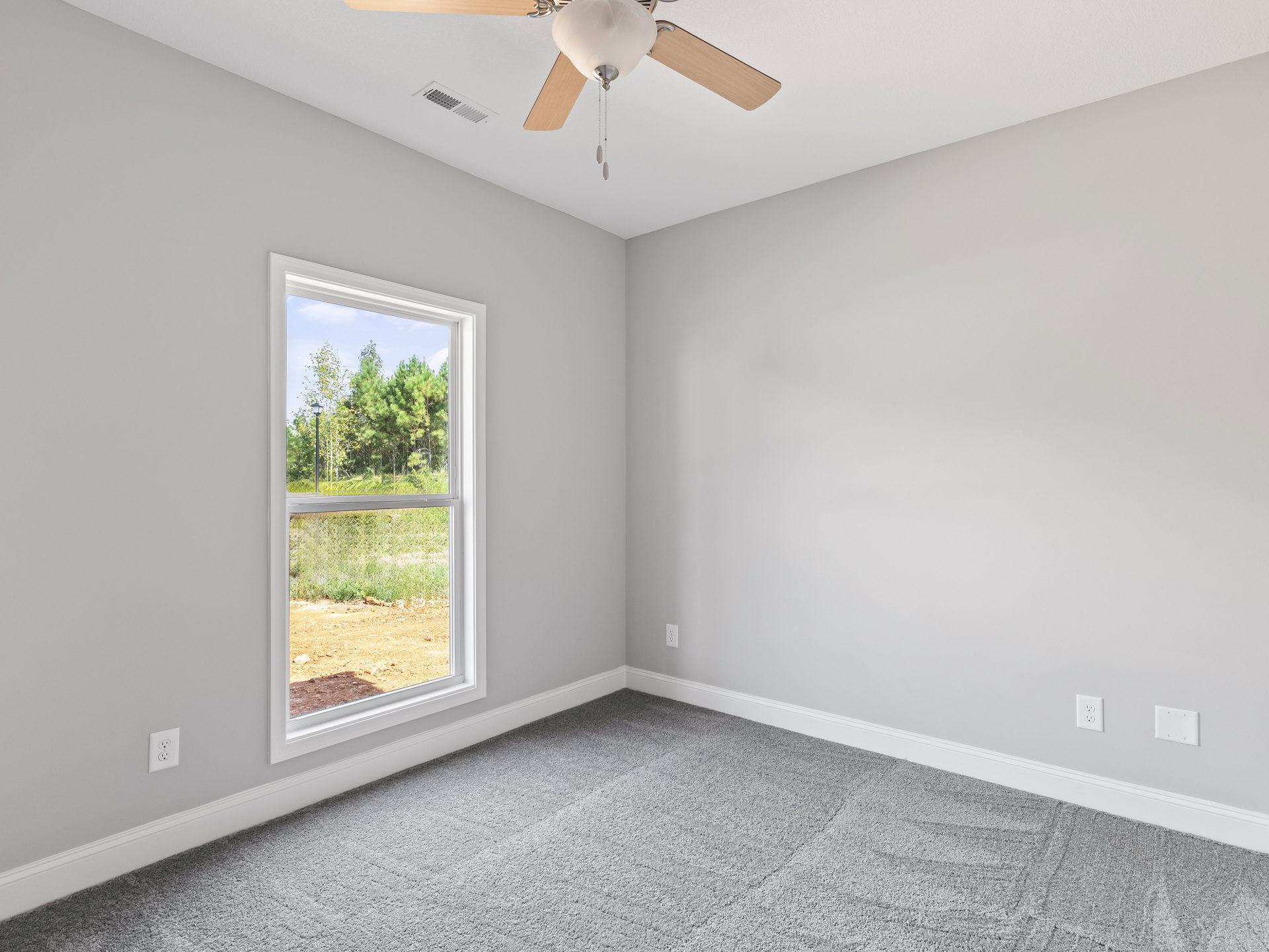 Bedroom with grey carpet flooring, white walls, ceiling fan, and window overlooking trees and a dirt field