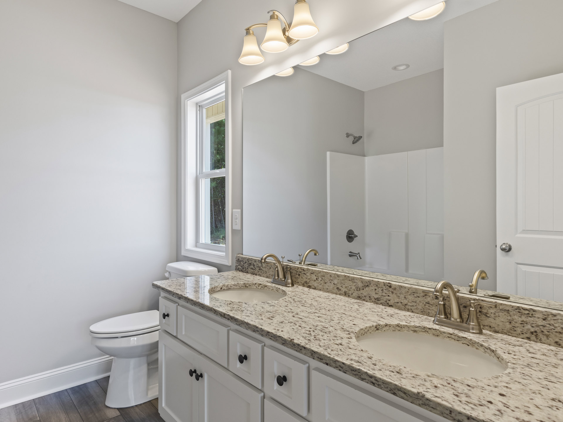 Bathroom with marble countertop, silver faucet, large wall mirror, closed toilet, white cabinetry, and window showing trees outside
