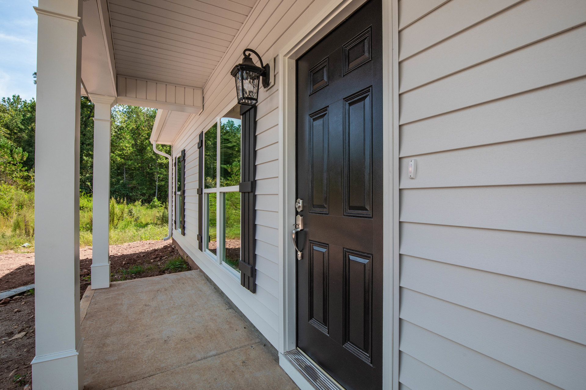 Black front door with modern handle on white siding, concrete walkway, porch light, and window adjacent to entry.