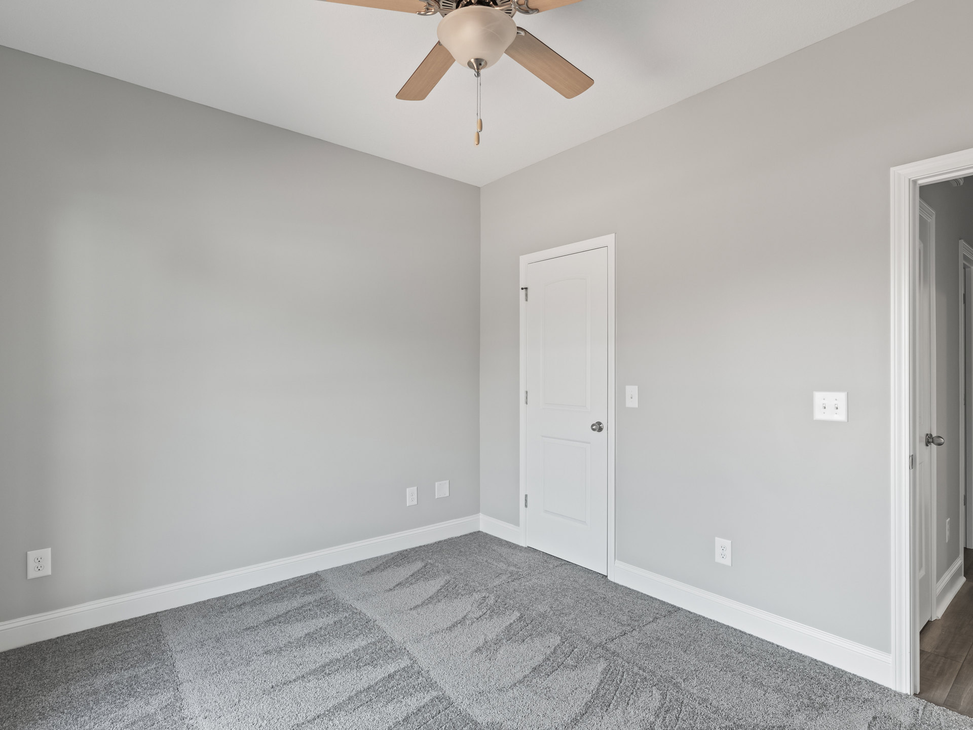 Ceiling fan with light fixture mounted on white plaster ceiling, carpeted floor, white door with silver door knob, light switch on wall.