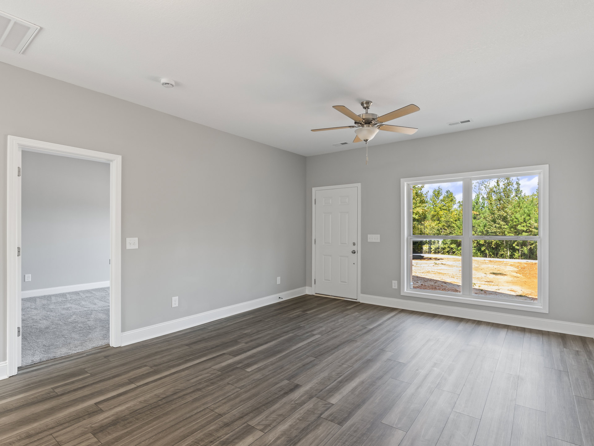 Ceiling fan with light fixture, wood laminate flooring, white walls, white door with silver handle, window overlooking trees