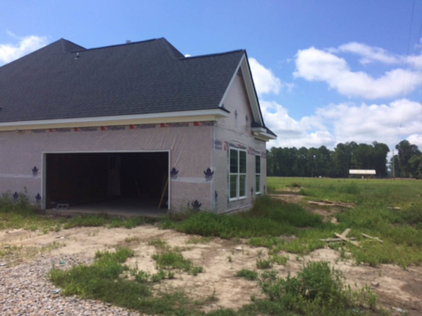 Partially built house with exposed framing, open garage, white-framed window, dirt patch with emerging grass, roof structure, grassy field and trees in background
