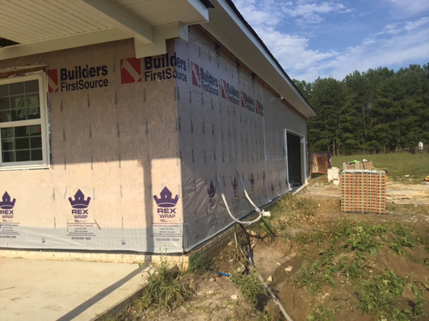 Modern house under construction with exterior walls wrapped in protective plastic sheeting, large windows, grassy lot, and surrounding trees under a partly cloudy blue sky