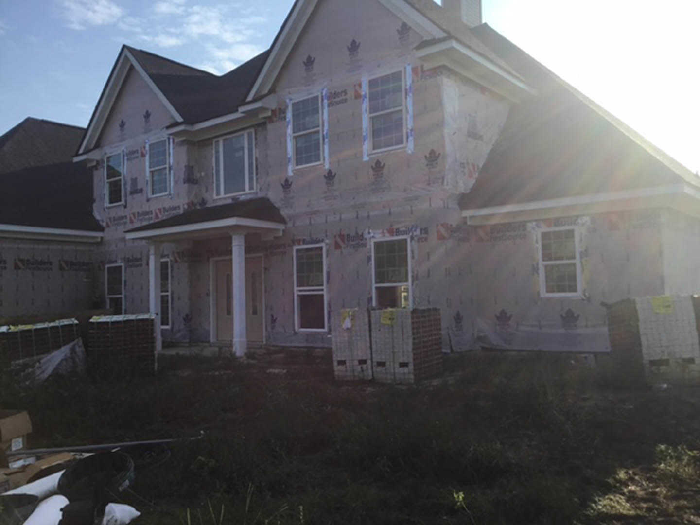 Two-story home under construction with exposed wooden framing, multiple square white-framed windows, unfinished siding, and a covered porch area against a cloudy sky