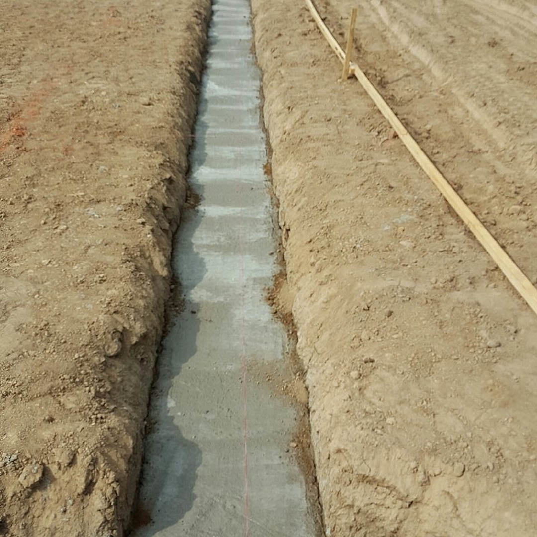 Deep trench dug into brown soil, bordered by a wooden fence, with rough dirt surfaces and scattered rocks visible.