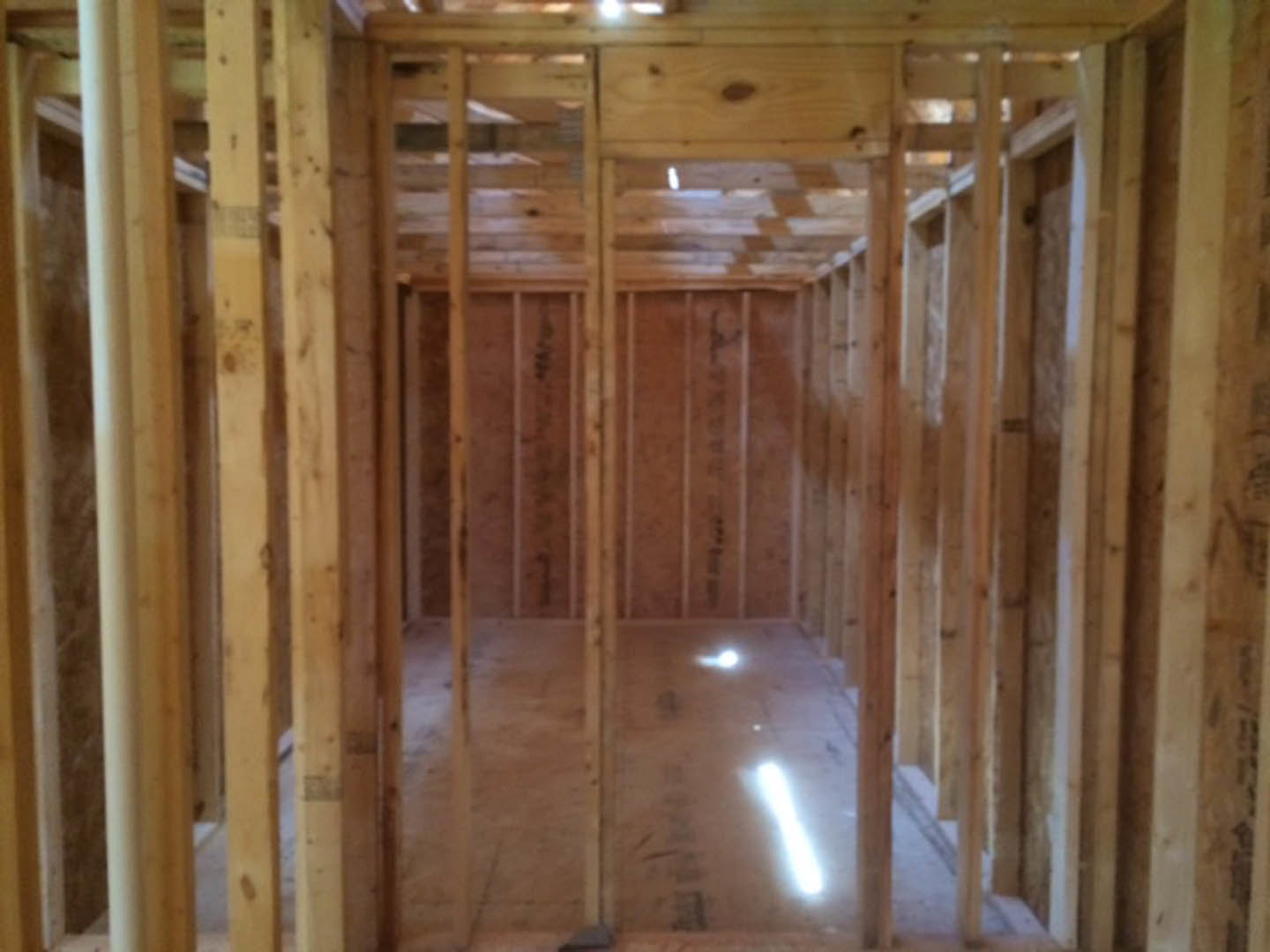 Room under construction with exposed wood framing, plywood walls, and beams; sunlight streaming through windows reflects off a tile floor.