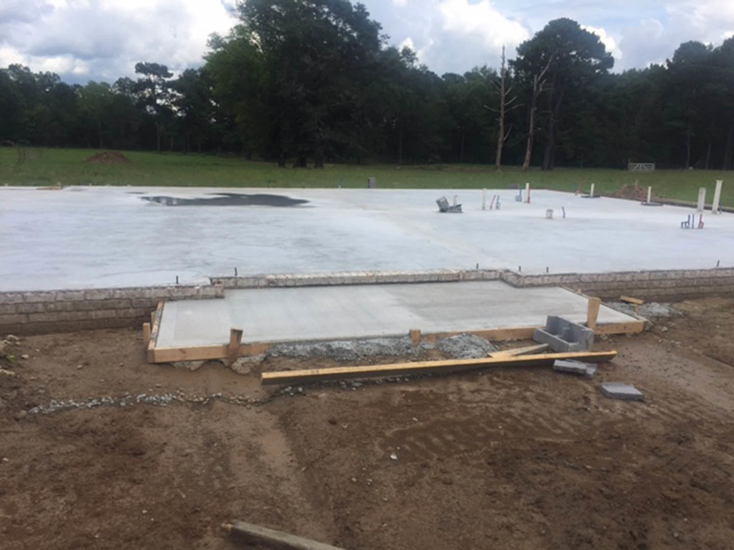Freshly poured concrete slab foundation surrounded by dirt field, wooden beams, and scattered construction materials, with trees and cloudy sky in the background