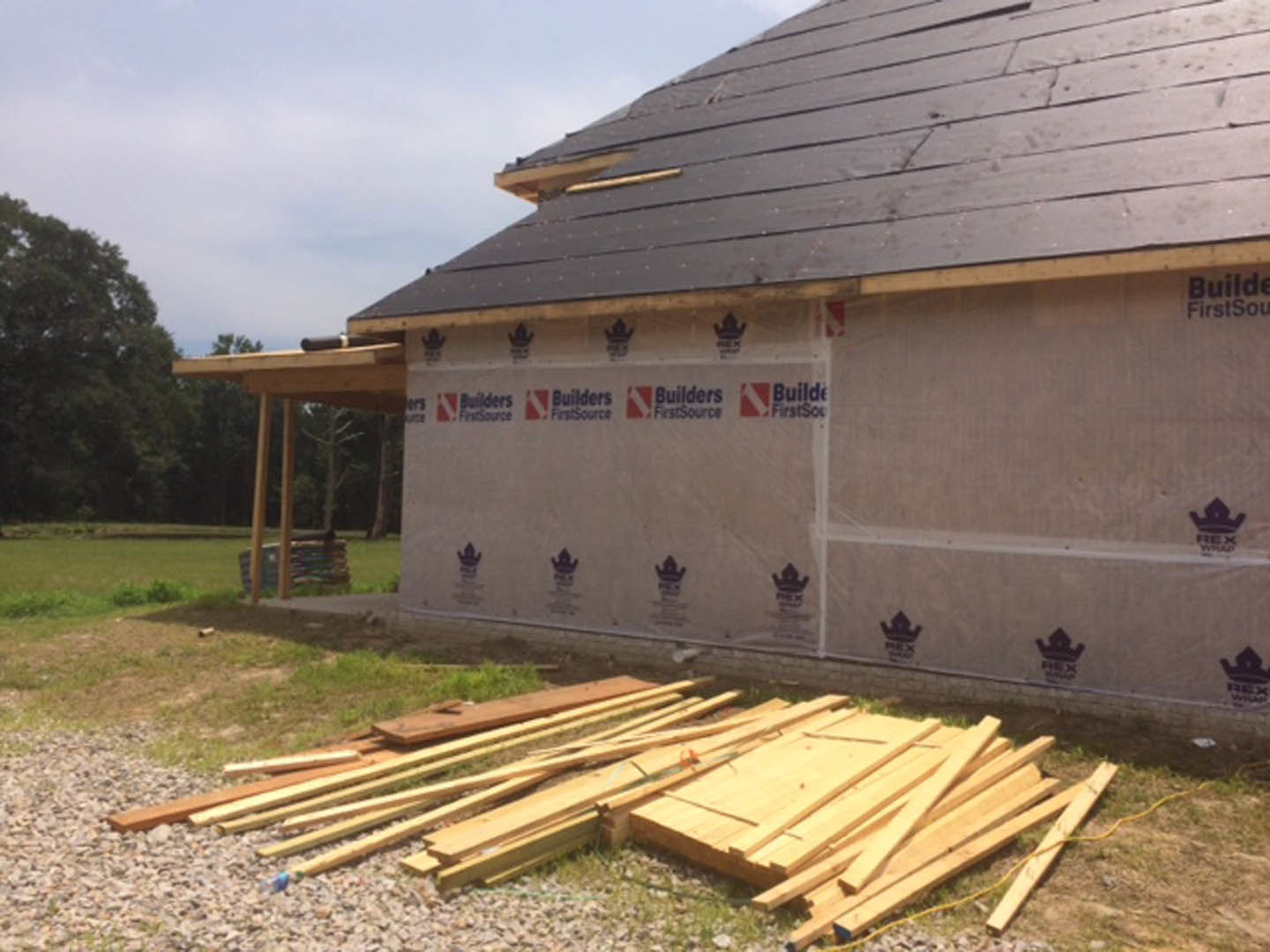 Modern home exterior under construction with horizontal wood plank siding, stacked lumber on grassy lot, and trees in background