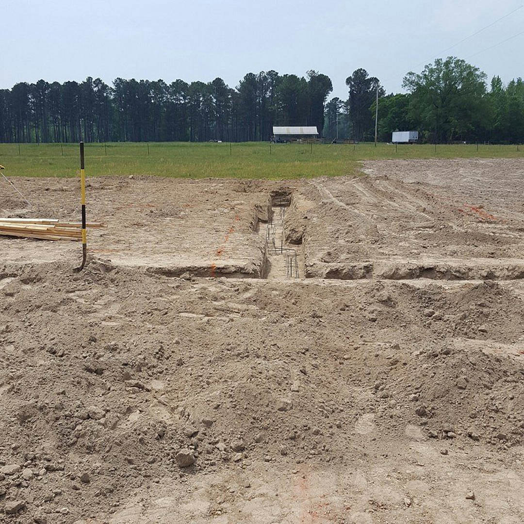 Excavated foundation pit surrounded by piles of wood, muddy soil, and grassy field, bordered by dense trees under a cloudy sky