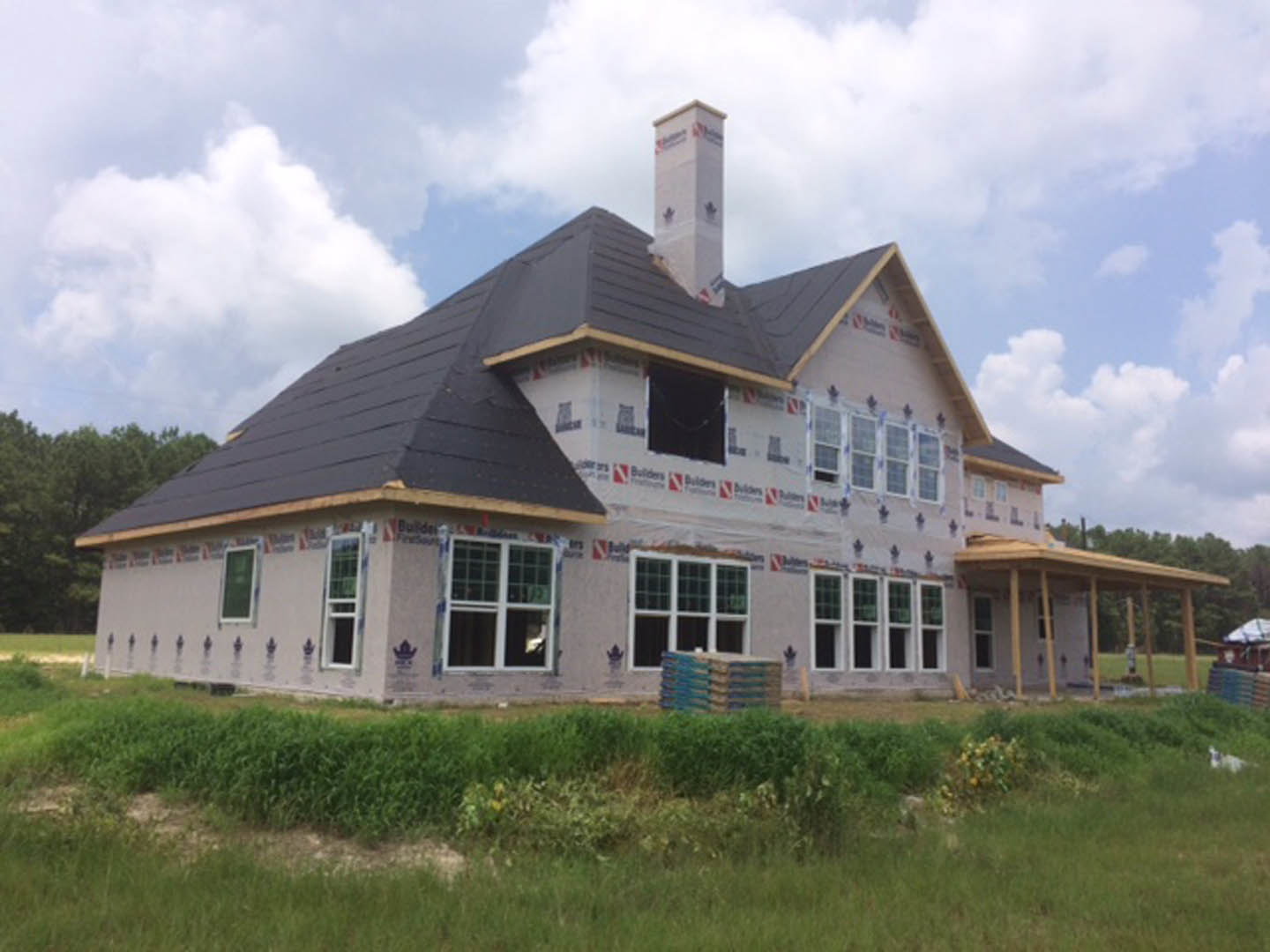 Partially built house with exposed framing, brick chimney, grassy yard, mature trees, covered porch, and large windows
