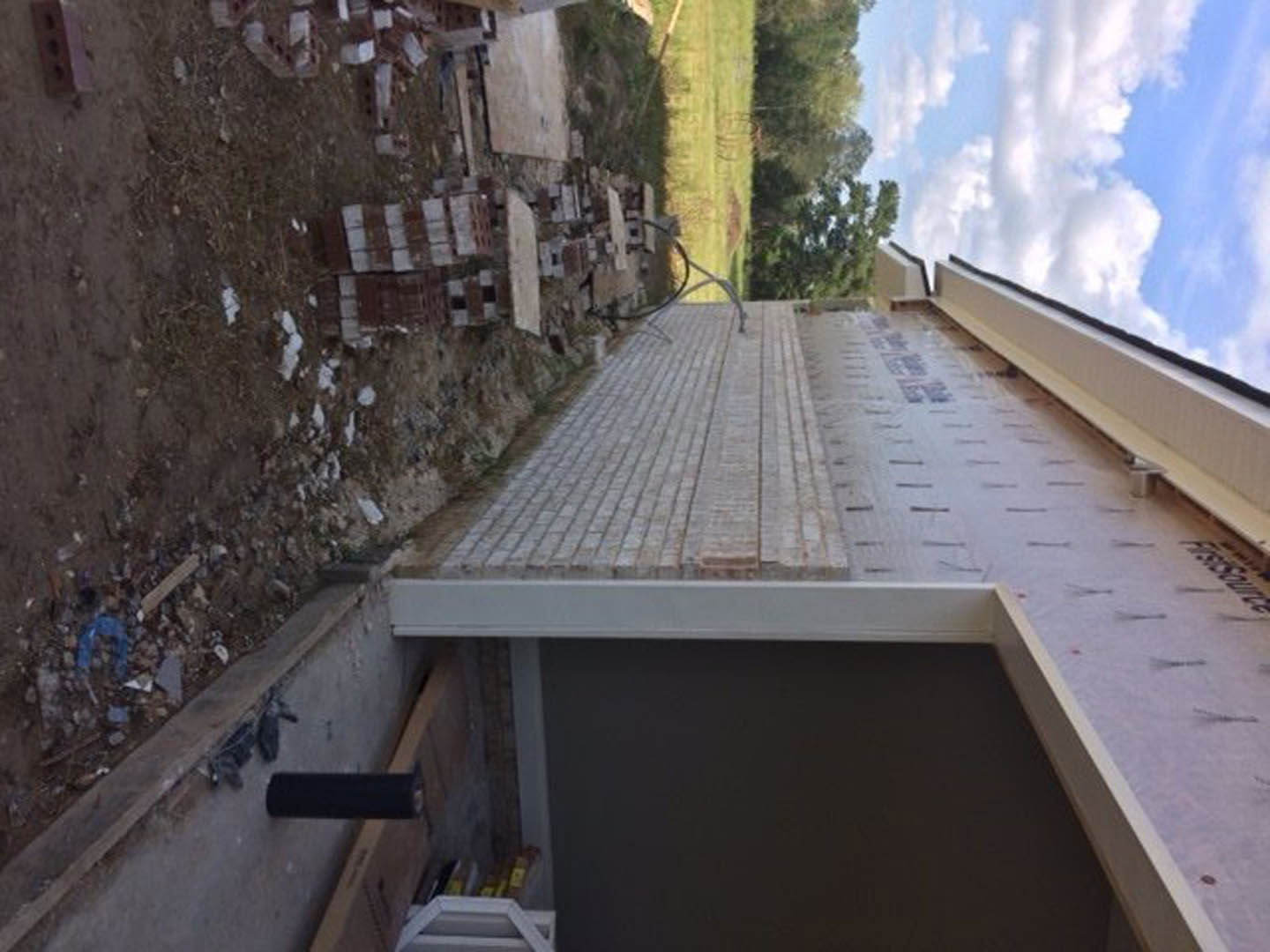 Wood-framed custom home under construction with exposed beams, piles of bricks and debris on dirt ground, blue sky and scattered clouds overhead, trees in background