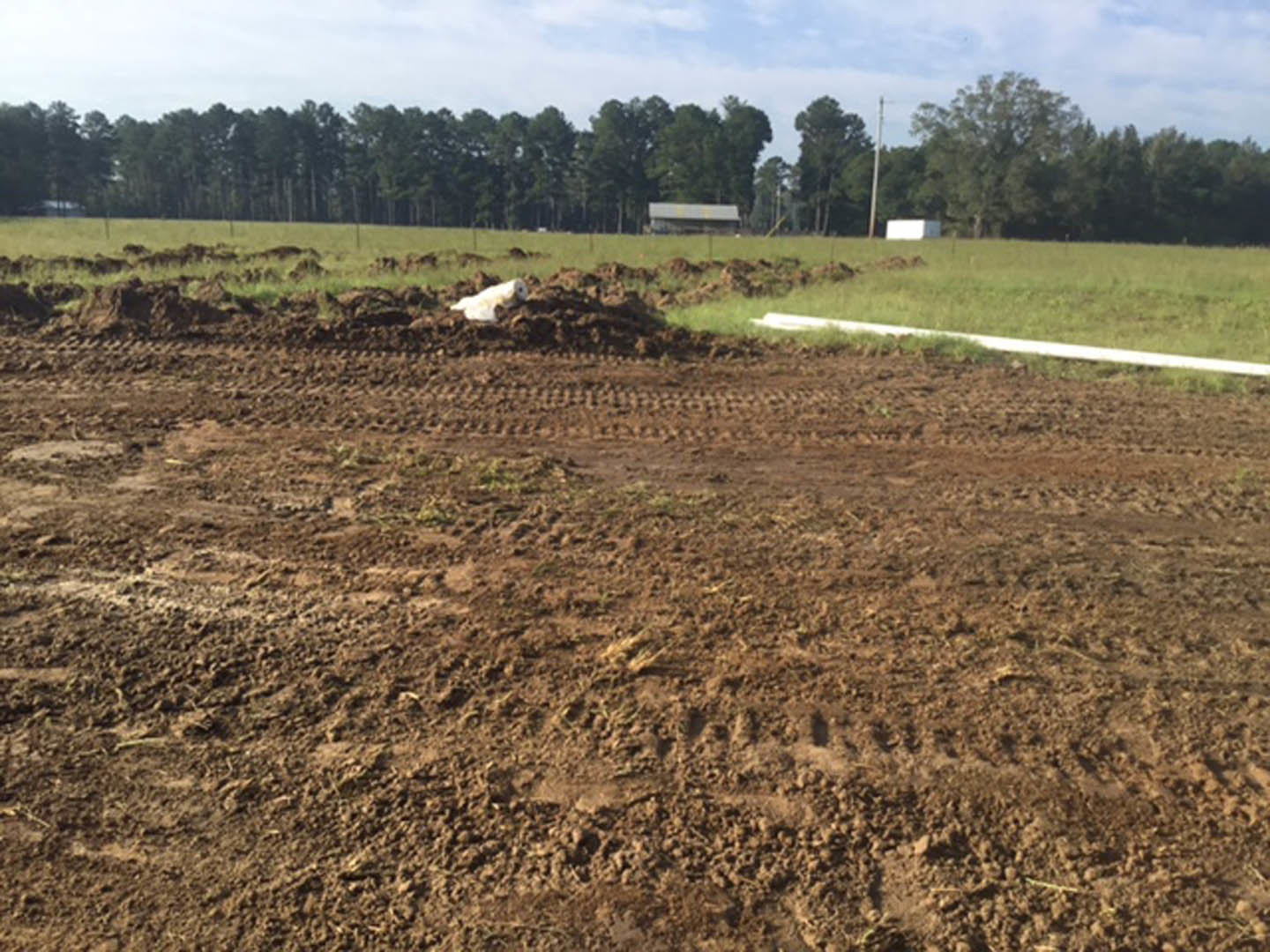 Dirt field bordered by green grass and scattered trees in the background under a partly cloudy sky