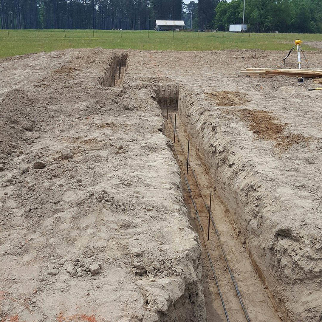 Dirt trench reinforced with metal rods, surrounded by grass and trees, surveyor's equipment nearby, building visible in background