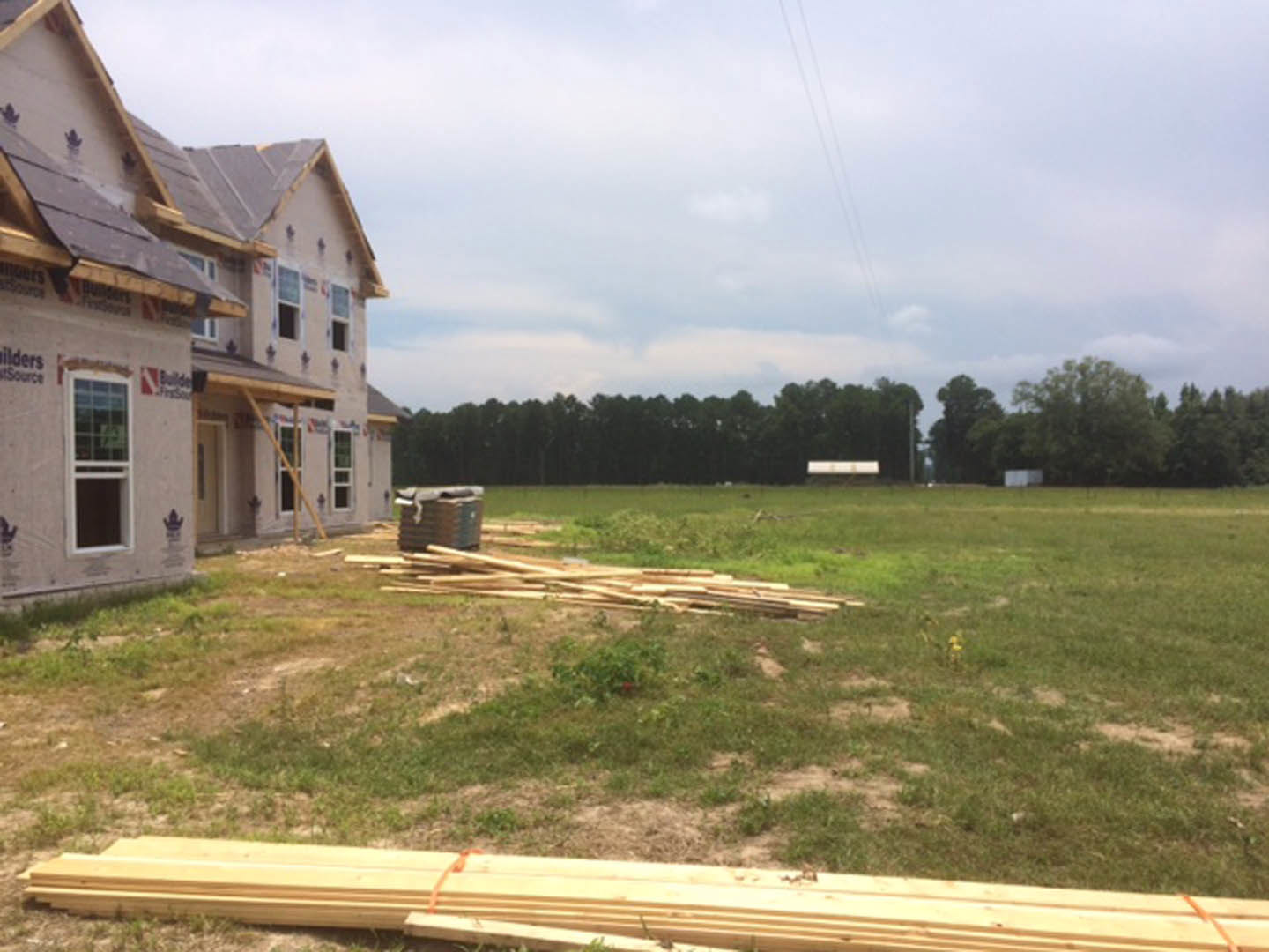 Two-story home with white-framed windows, expansive grassy yard, scattered wood planks and lumber, trees and clouds in the background