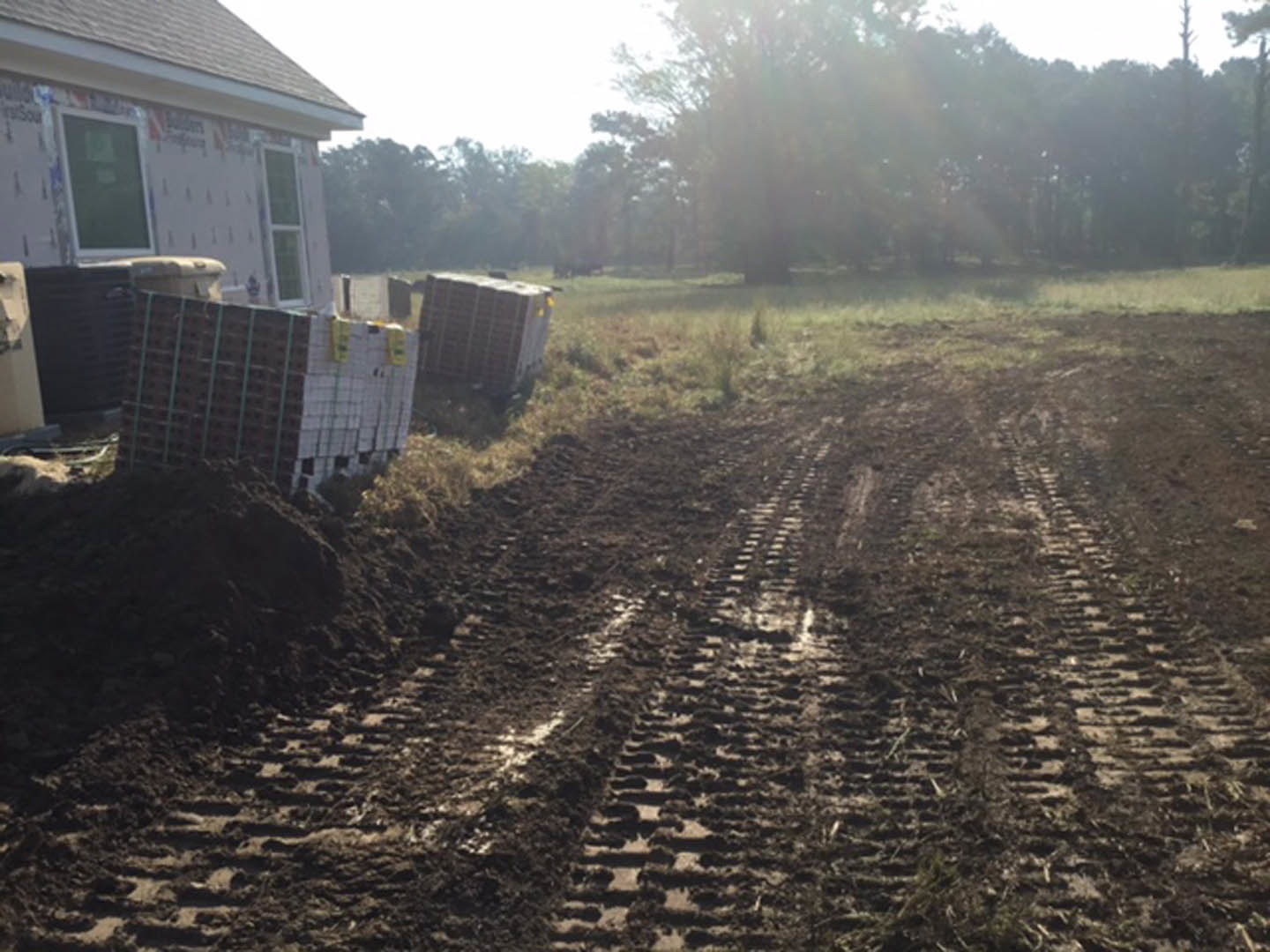 Dirt road with deep tire tracks leading to a grassy land lot, surrounded by trees and soil, under an open sky