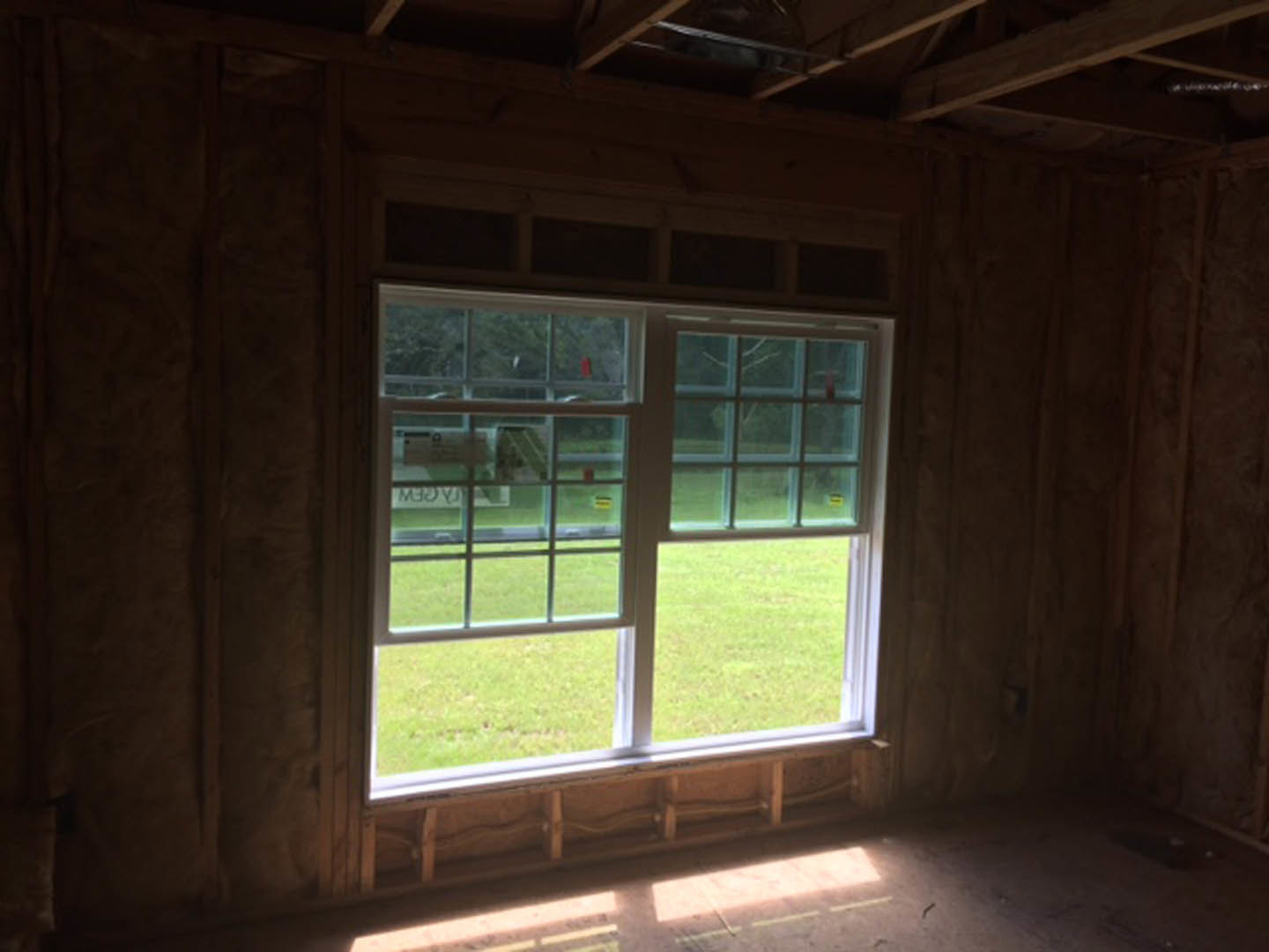 Large window framed by dark hardwood beams, sunlight illuminating wood plank flooring, grassy field visible outside