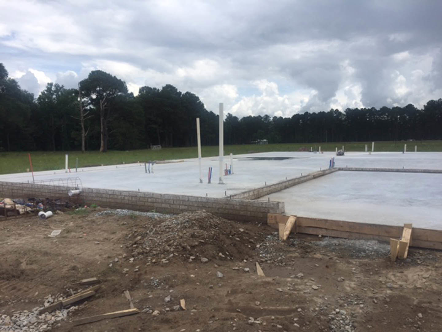 Concrete foundation with white poles set in a grassy field, piles of dirt and rocks nearby, cloudy sky overhead, trees in the background.