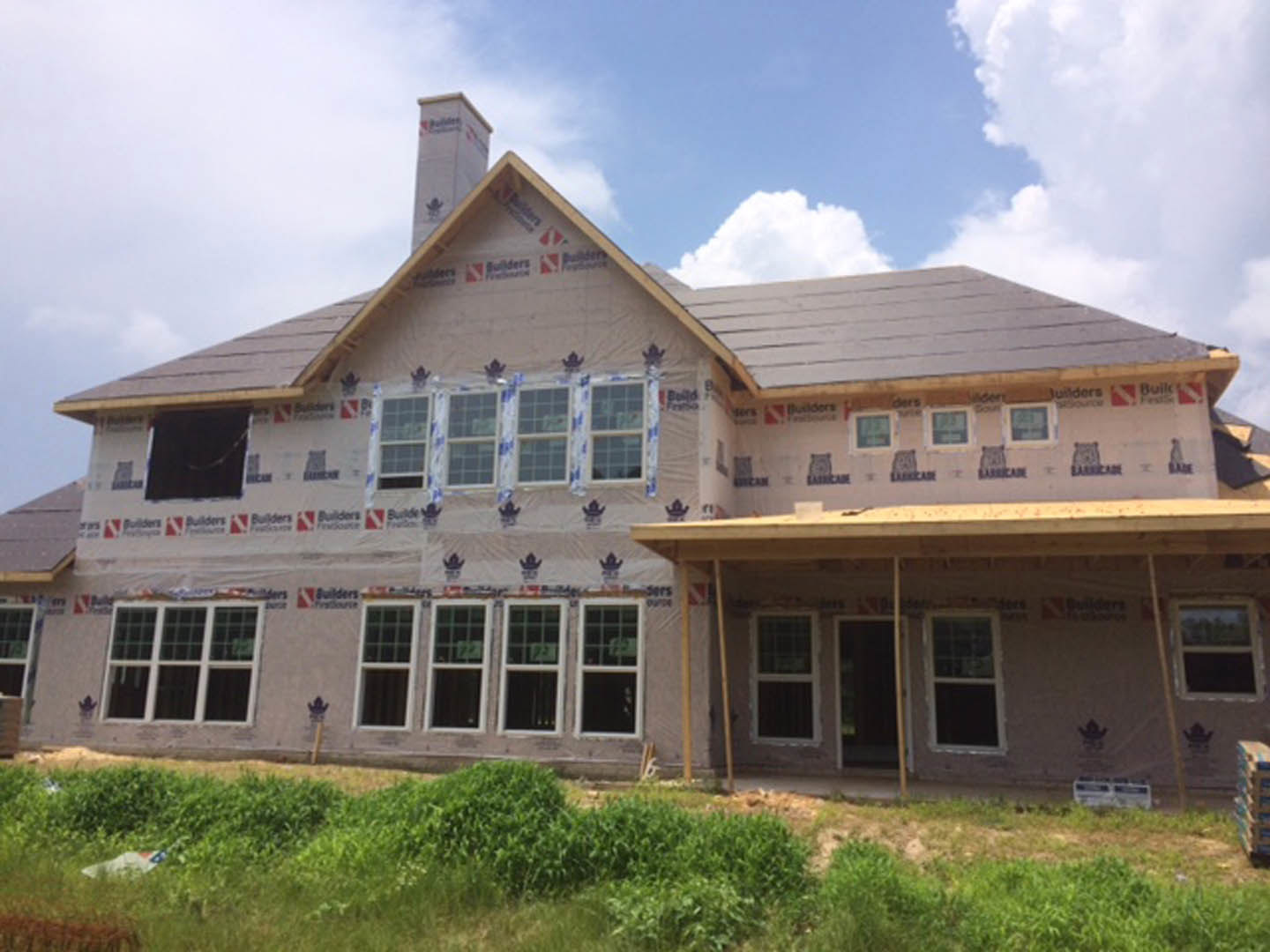 Partially built house with exposed framing, covered porch, green lawn, and blue sky in background