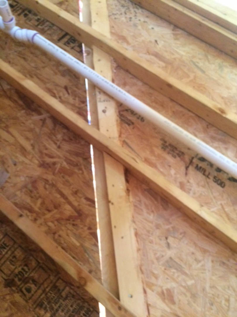 White PVC pipe running through unfinished plywood and hardwood framing in a residential interior construction.
