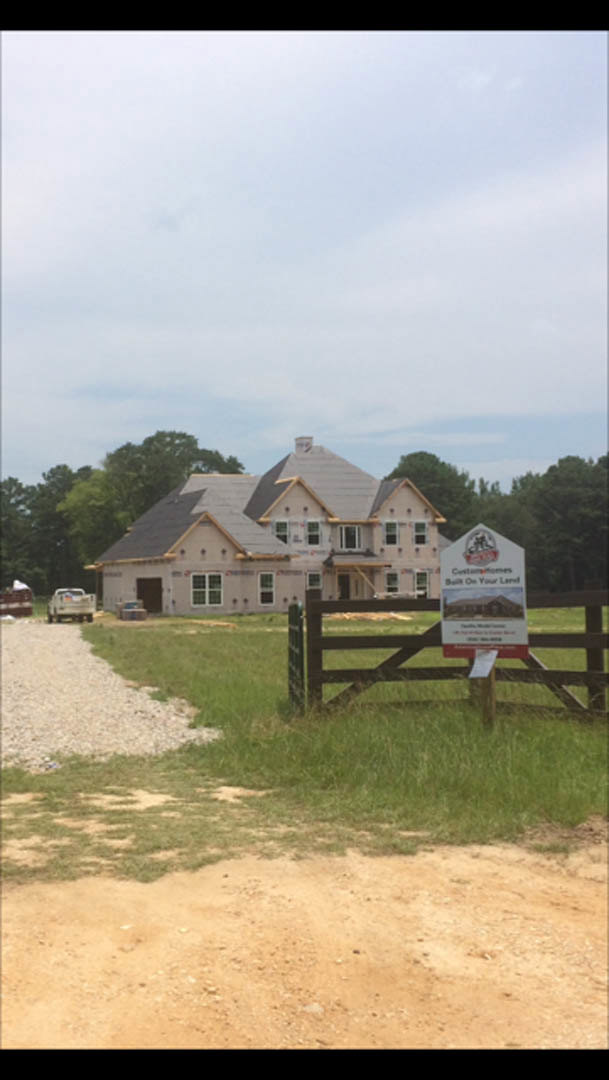 Wood-framed house under construction behind chain-link fence with contractor sign, surrounded by gravel, grass, and dirt field, few windows installed, cloudy sky overhead.