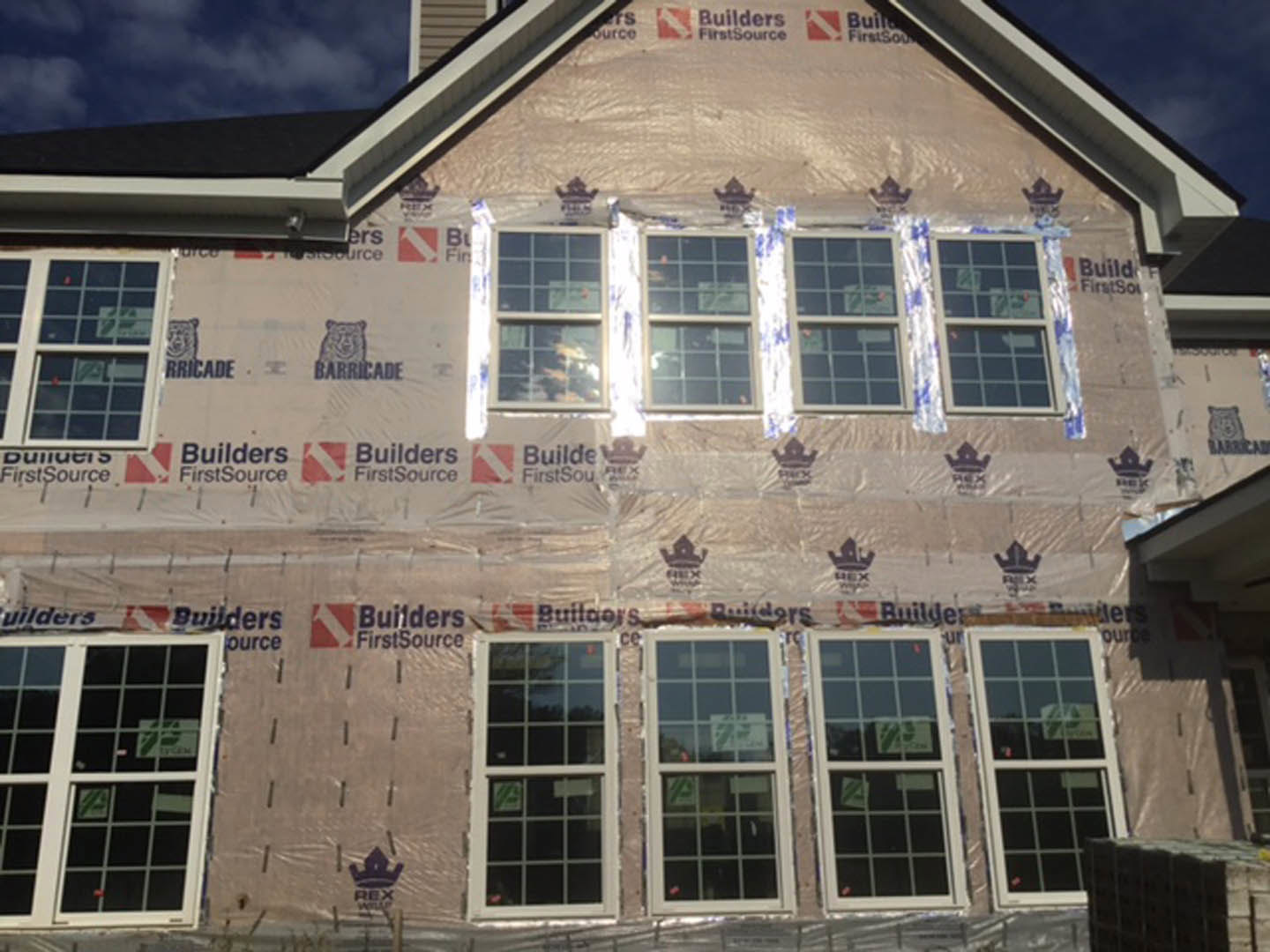 White multi-pane window with plastic sheeting taped over the frame, exterior wall partially finished, construction sign visible on glass