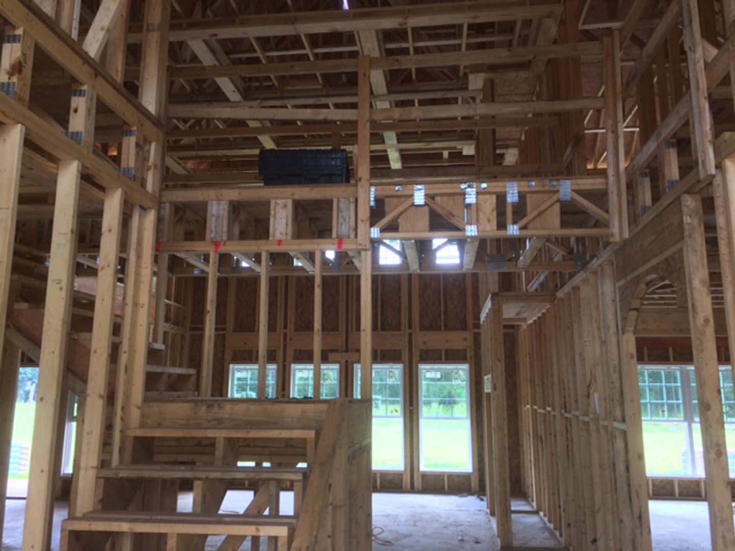 Exposed wood framing and beams in a partially constructed interior, with a close-up of a wooden staircase and a window overlooking a fenced yard with trees.