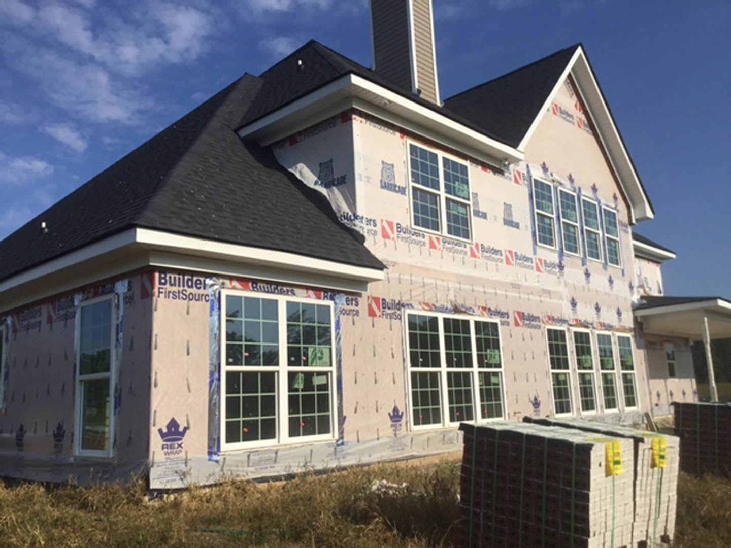 Partially built house with exposed framing, stacks of pallets and bricks in front, multiple sash windows installed, white siding visible, cloudy sky overhead
