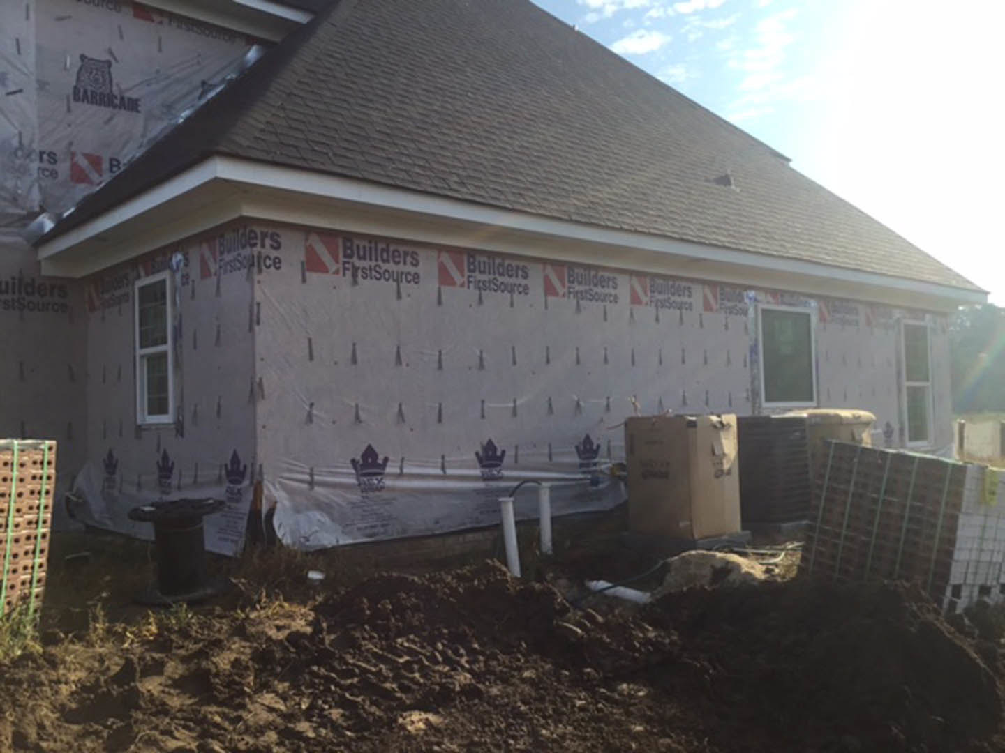 Partially built home with exposed framing, unfinished exterior walls, pile of dirt in front yard, construction bucket, visible window opening, and scattered building materials.