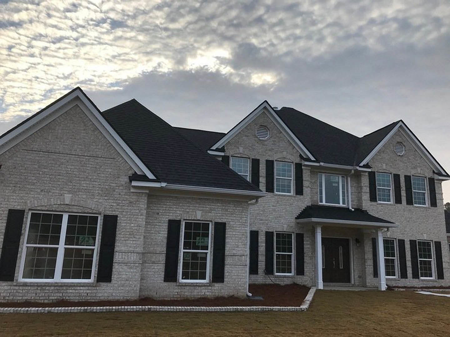 Two-story house with black roof, gray siding, covered porch, large windows, green lawn, and overcast sky