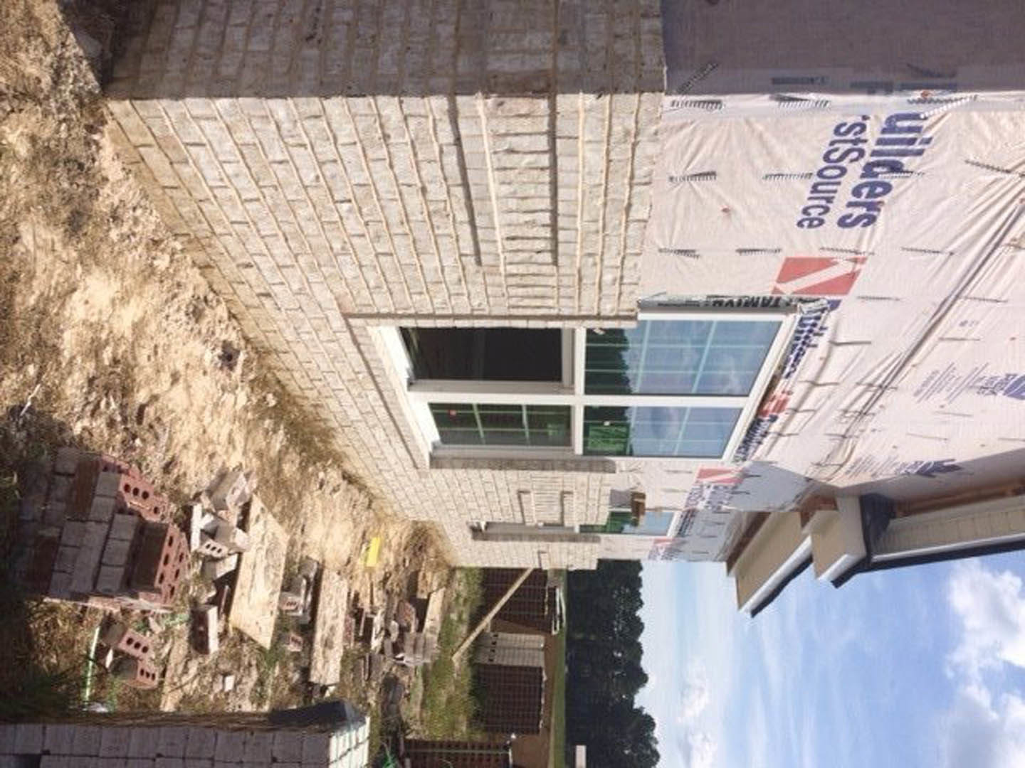 Partially built brick exterior wall with exposed mortar, window opening framed, construction materials visible, cloudy sky in background