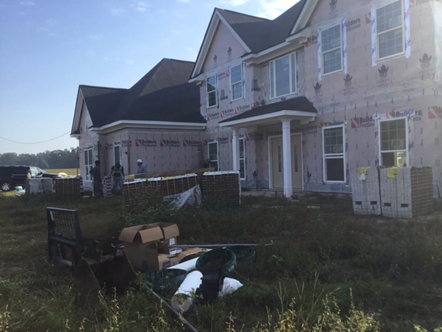 Two-story cottage-style home with white siding, large windows, and gabled roof, surrounded by green grass and plants, with several people standing near the front entrance.