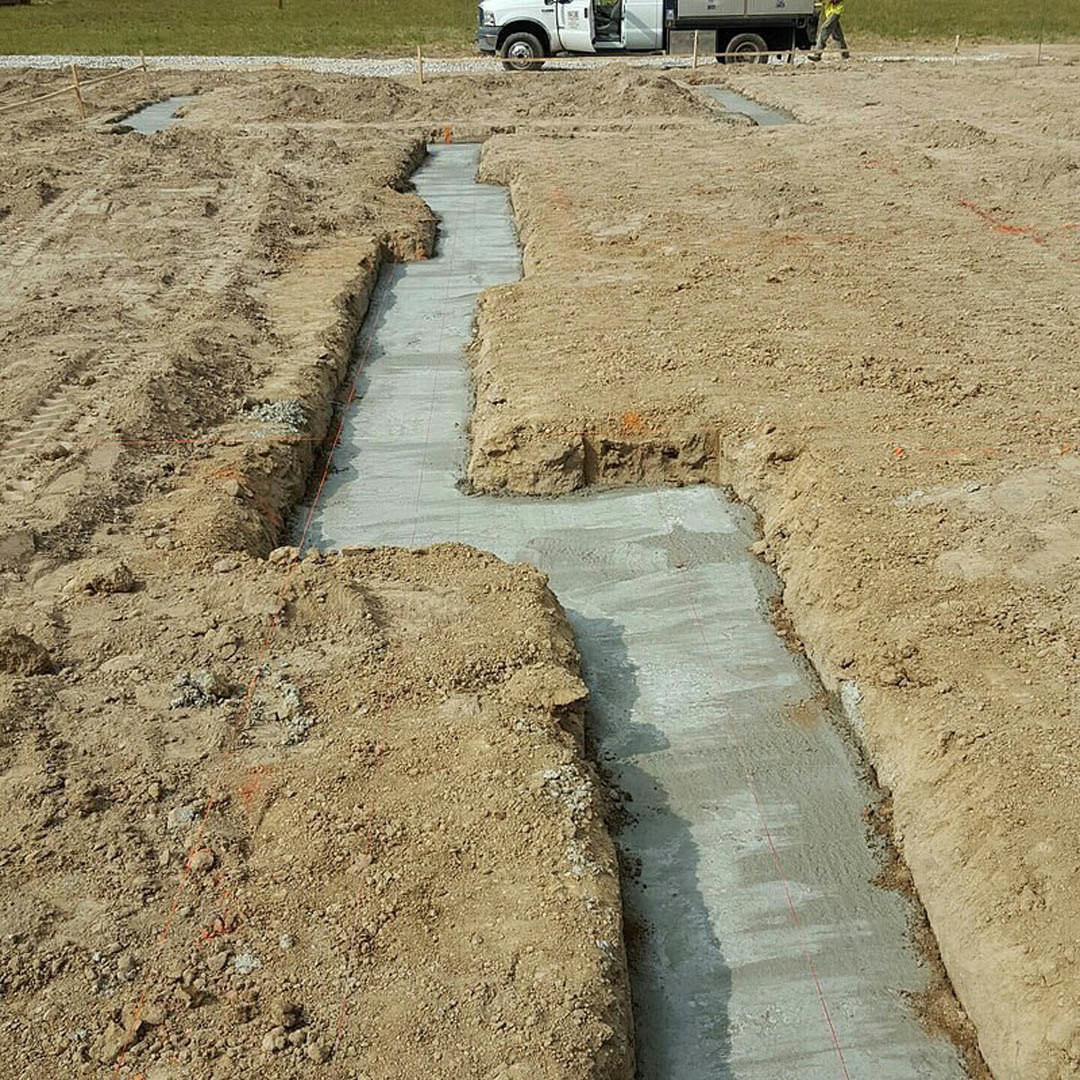 White work truck parked on dirt construction site beside grassy field, concrete trench and small stream visible, person in yellow shirt standing nearby, scattered rocks on ground.
