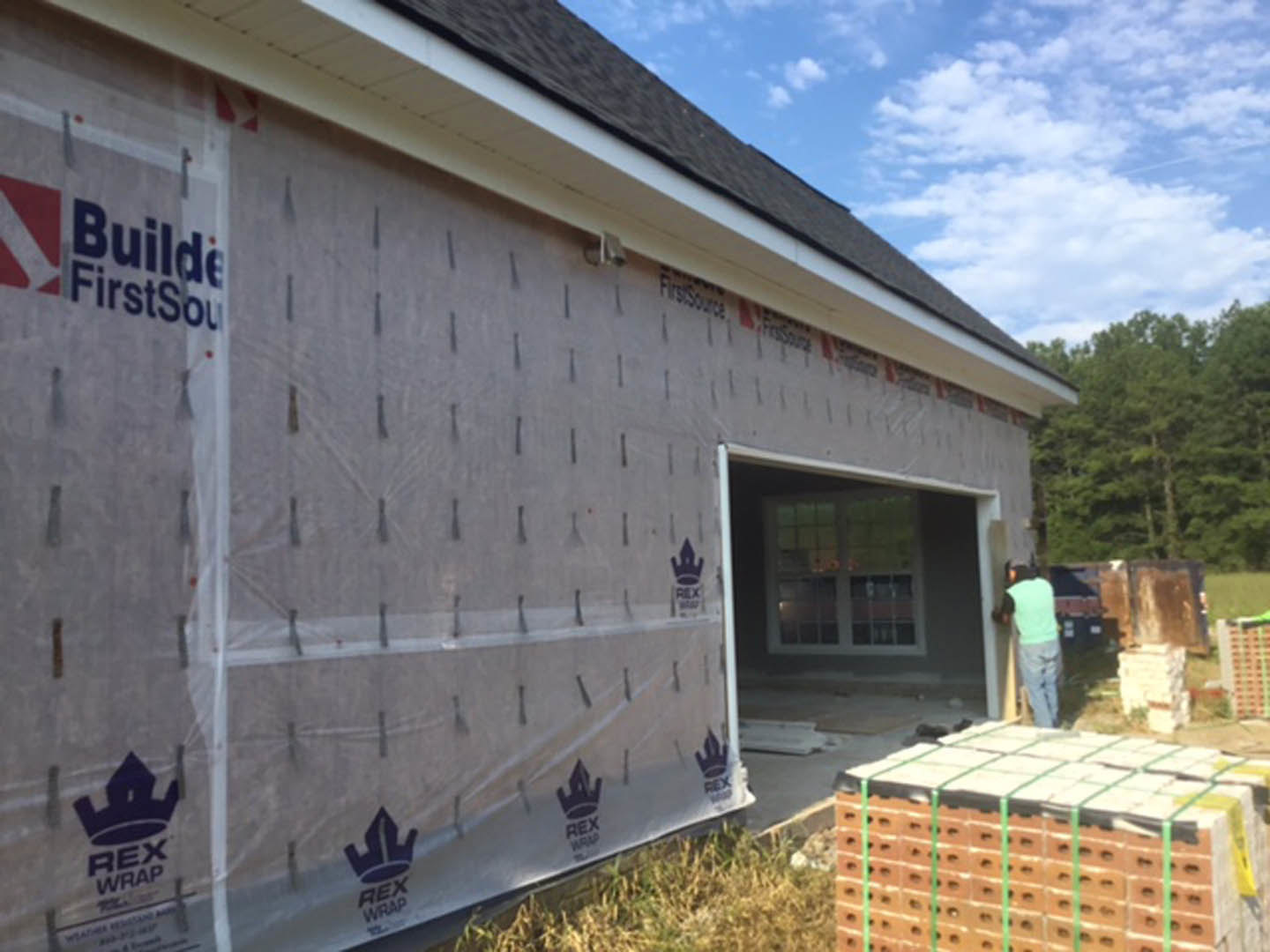 Modern home under construction with plastic sheeting covering windows, stack of bricks secured with green strap in foreground, man in green shirt standing near entrance, trees and