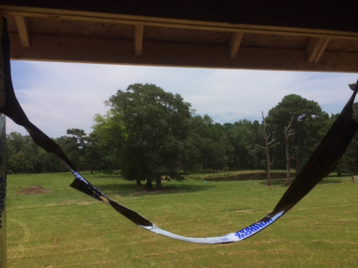 Hammock suspended between trees in a grassy backyard, visible tape wrapped around hammock fabric, open sky and field in background