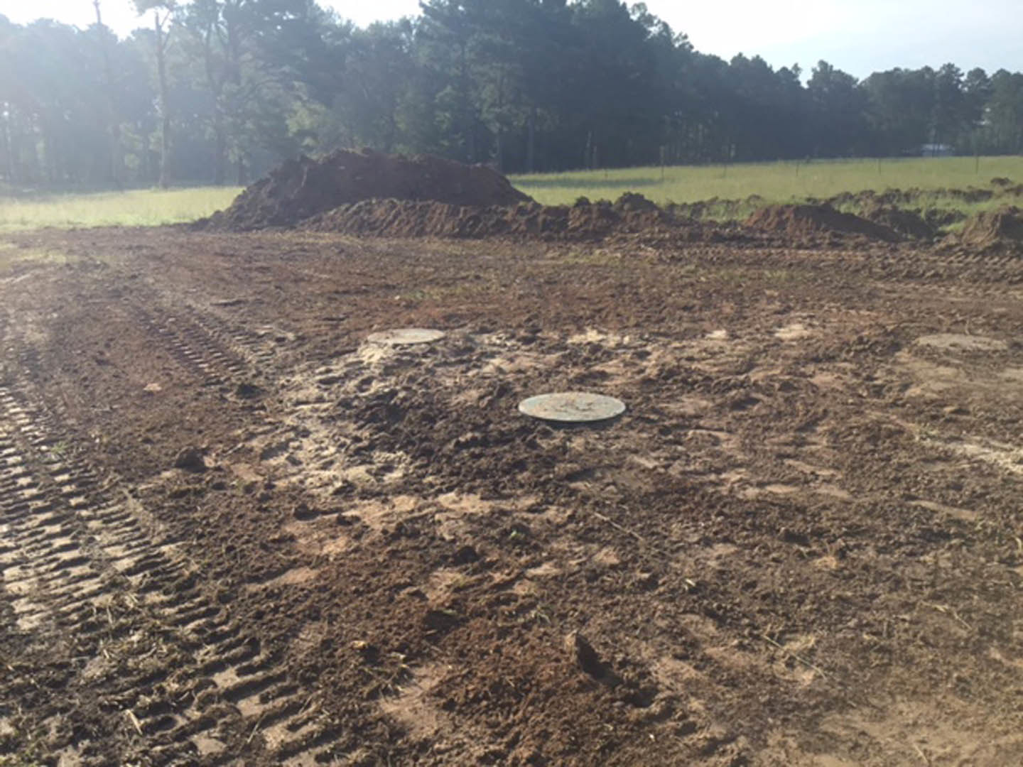 Dirt field with visible tire tracks, scattered grass, a metal manhole cover, and trees in the background