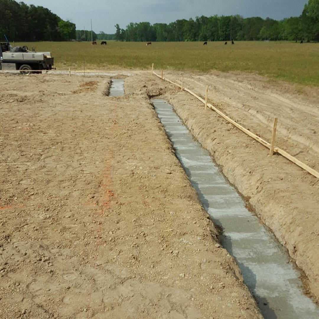 Wide dirt trench cut through grassy field with scattered trees in background, pickup truck parked beside excavation, exposed soil and rough ground surface visible under open sky.