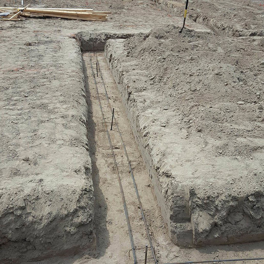 Wide dirt trench with rough earth sides, large pile of soil nearby, scattered wood planks on ground, close-up of yellow and black handled tool, outdoor construction site with