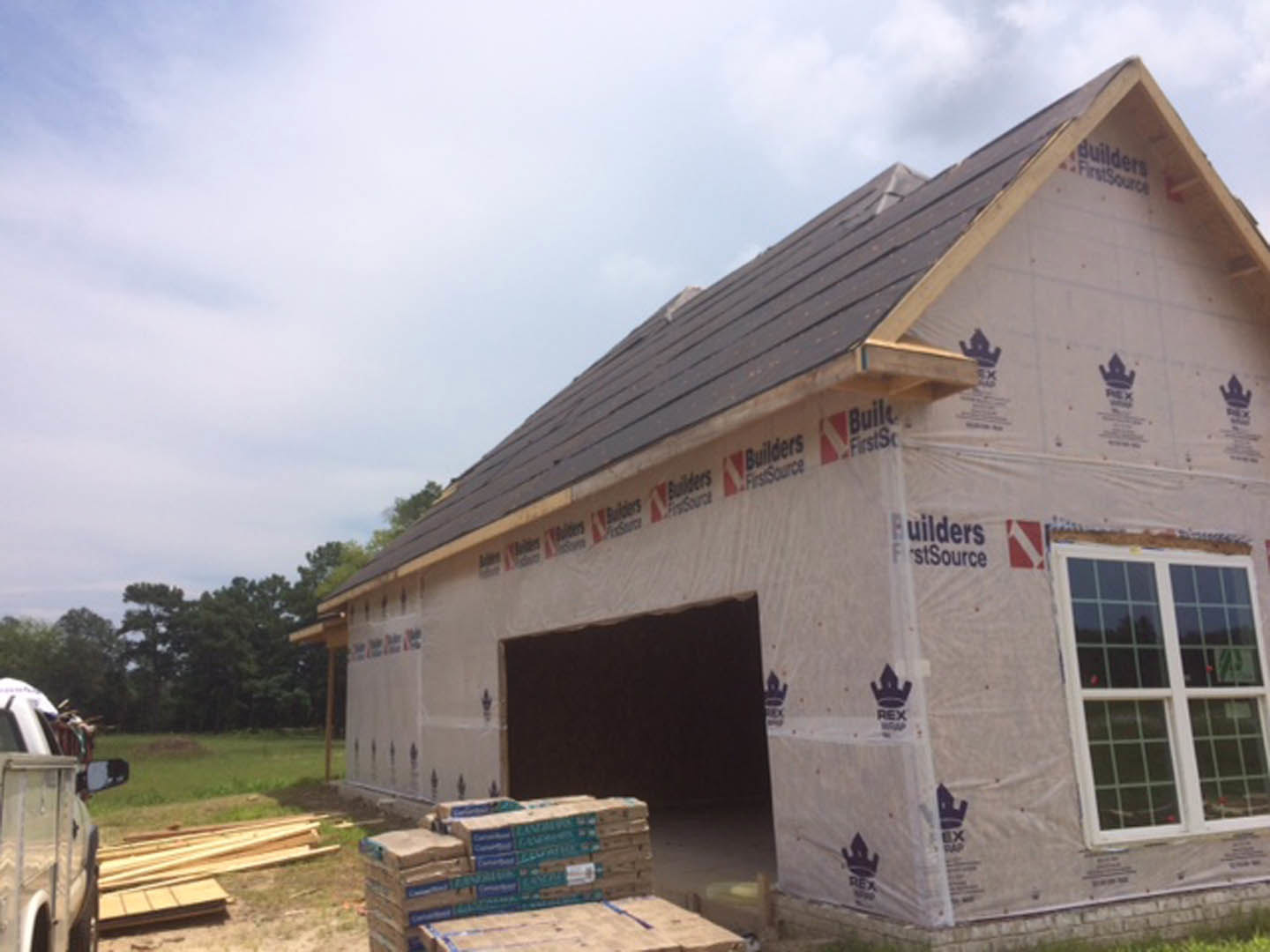 Framed house under construction with exposed lumber, large attached garage, unfinished windows, grassy yard, and scattered building materials