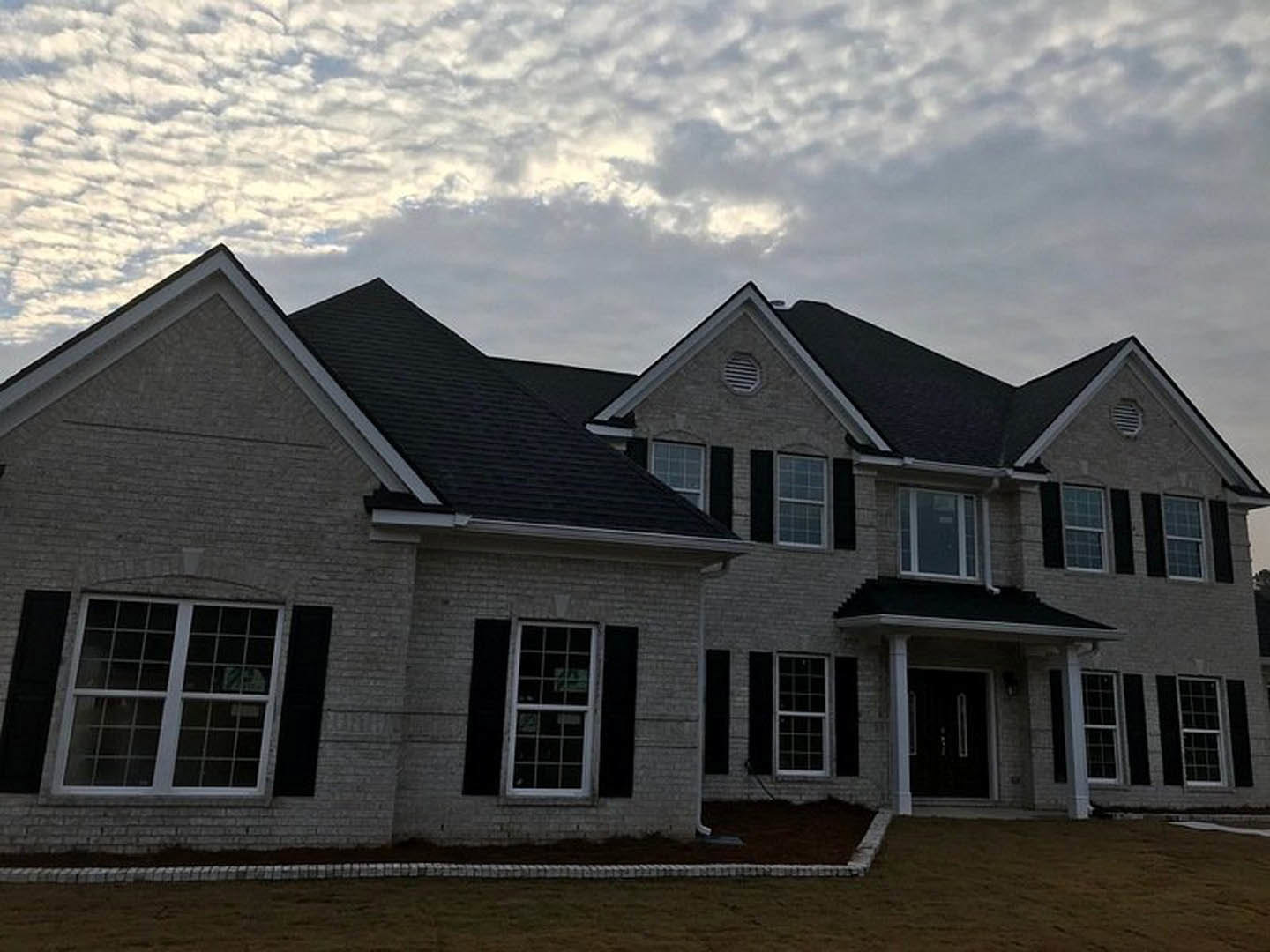 Two-story house with light siding, multi-pane windows, front lawn, paved driveway, and overcast sky with dense clouds