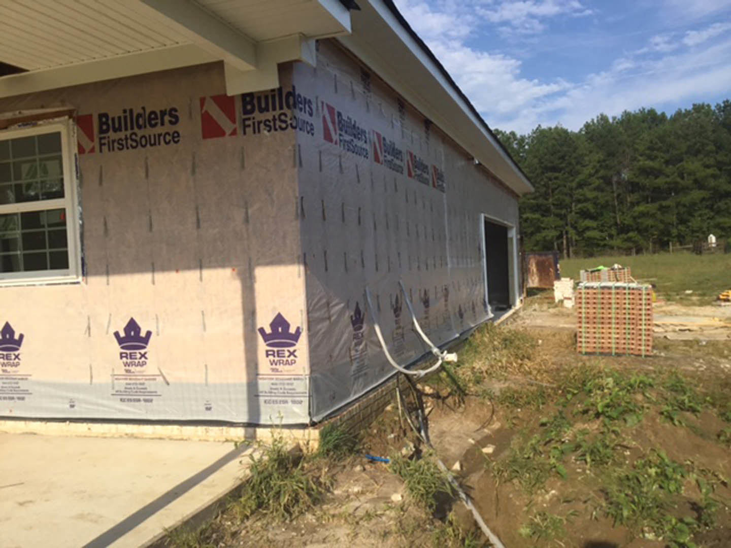 House under construction with plastic sheeting covering exterior walls, exposed window frame, grassy yard, and surrounding trees under partly cloudy sky